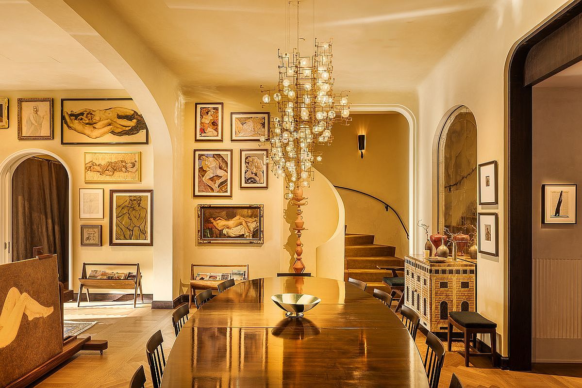 This is an elegant dining room featuring a long, polished wooden table surrounded by chairs, illuminated by a striking, multi-bulb chandelier. The walls are adorned with numerous framed artworks, creating a gallery-like atmosphere, and an arched doorway leads to a staircase. The room exudes a sense of classic sophistication and artistic flair.