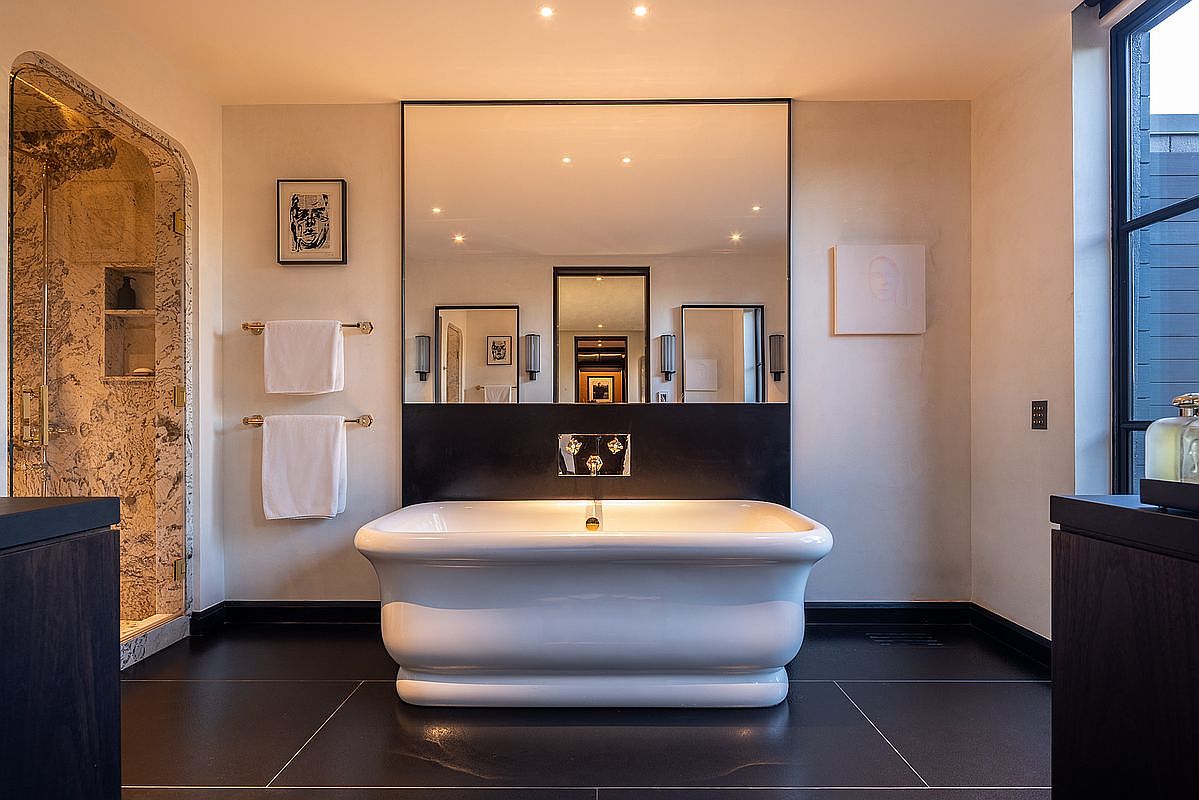 This is a luxurious primary bathroom featuring a freestanding white bathtub as the focal point, set against a backdrop of a large mirror and dark paneling. The room is illuminated by recessed lighting, and the dark tile flooring adds a touch of sophistication. A glass-enclosed shower is visible to the left, and the overall impression is one of modern elegance and relaxation.