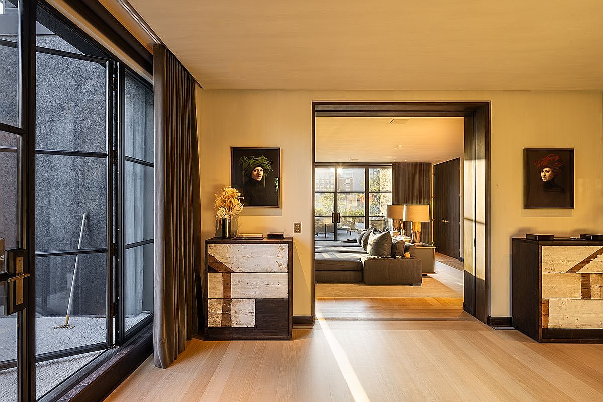 This interior shot showcases a stylish living room with hardwood floors and a neutral color palette. The room features a large window with black frames, a comfortable gray sectional sofa, and decorative artwork on the walls. A doorway leads to another room, creating an open and inviting atmosphere.