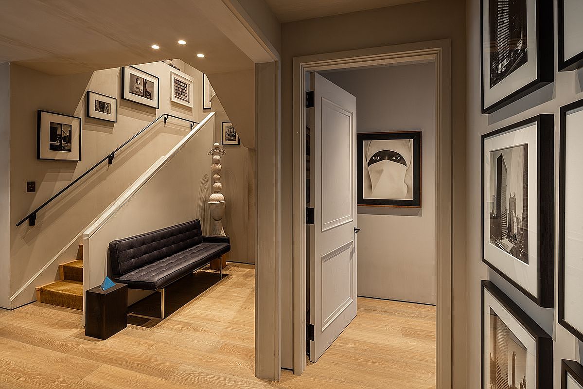 This interior shot showcases a well-lit hallway and staircase area, featuring light wood flooring and neutral-toned walls adorned with framed artwork. A modern bench sits beneath the staircase, adding a touch of contemporary style, while an open doorway reveals a glimpse into another room, enhancing the sense of depth and space. The overall impression is clean, sophisticated, and inviting.