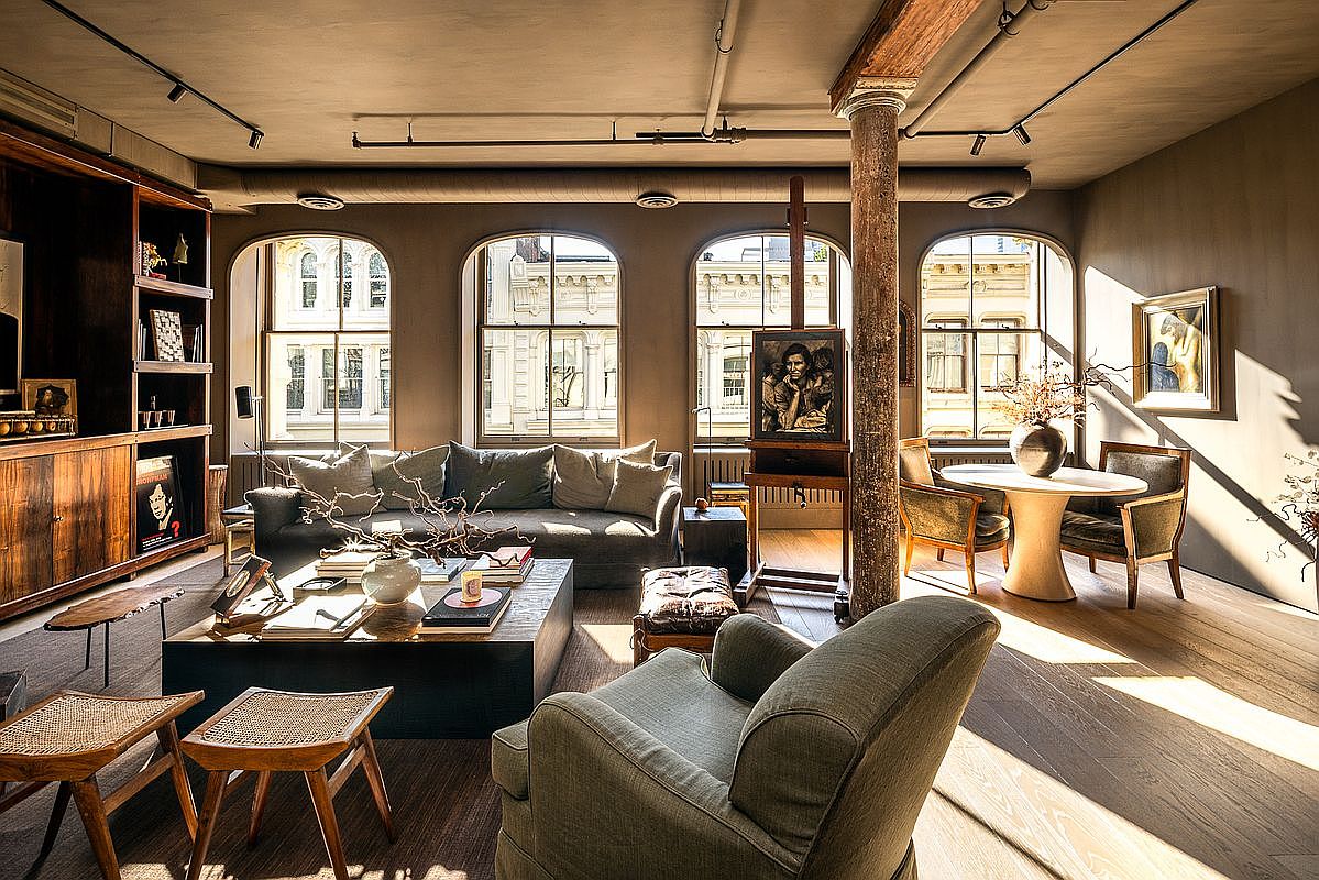 This is a beautifully designed living room featuring a blend of modern and classic elements. The room is bathed in natural light from large arched windows, highlighting the hardwood floors and comfortable seating arrangement. A large coffee table is adorned with books and decorative items, while an easel with a painting adds an artistic touch to the space.
