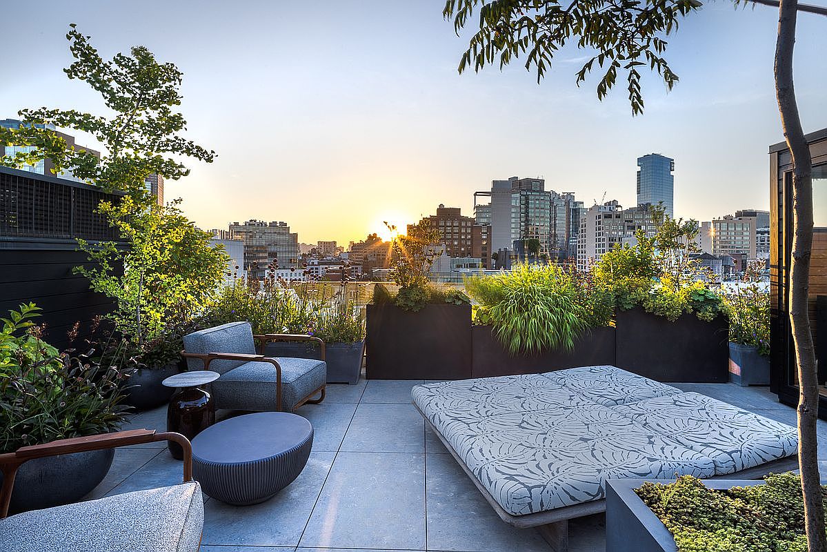 This image showcases a luxurious rooftop patio with stunning city views at sunset. The space is furnished with comfortable seating, including armchairs and a daybed, surrounded by lush greenery in modern planters. The overall impression is one of sophisticated urban living and relaxation.
