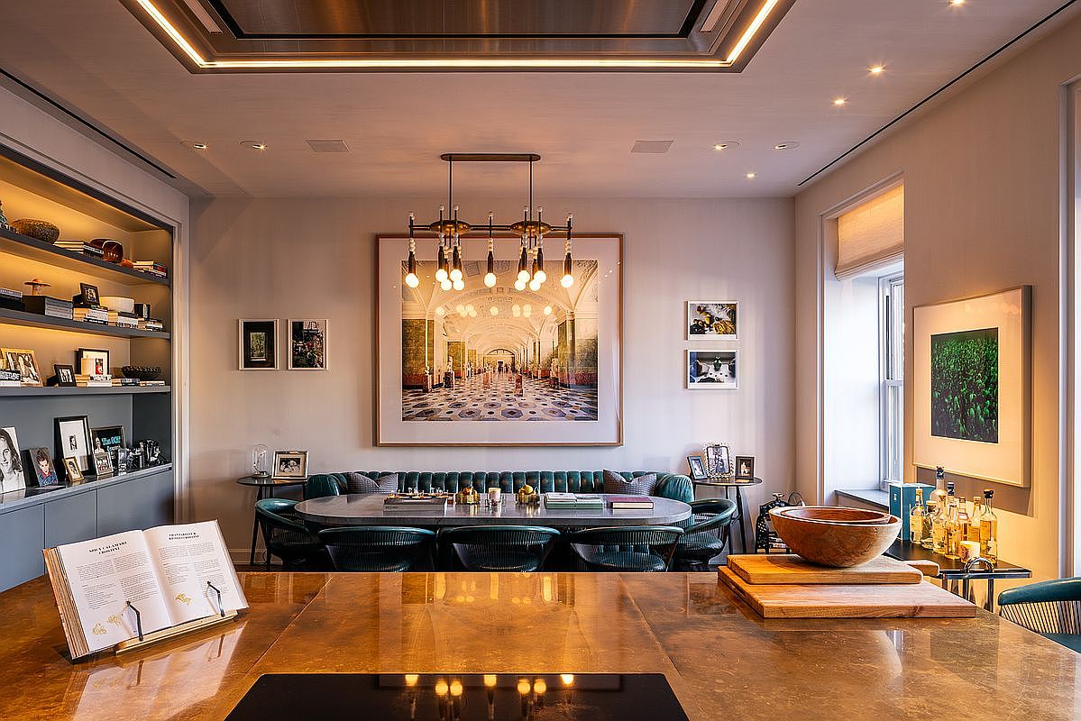 This interior shot showcases a sophisticated dining area with a large, oval table surrounded by green chairs and a tufted green banquette. A modern chandelier hangs above the table, and a large framed artwork adorns the wall, adding a touch of elegance. The room is well-lit with recessed lighting and natural light from a nearby window, creating a warm and inviting atmosphere.