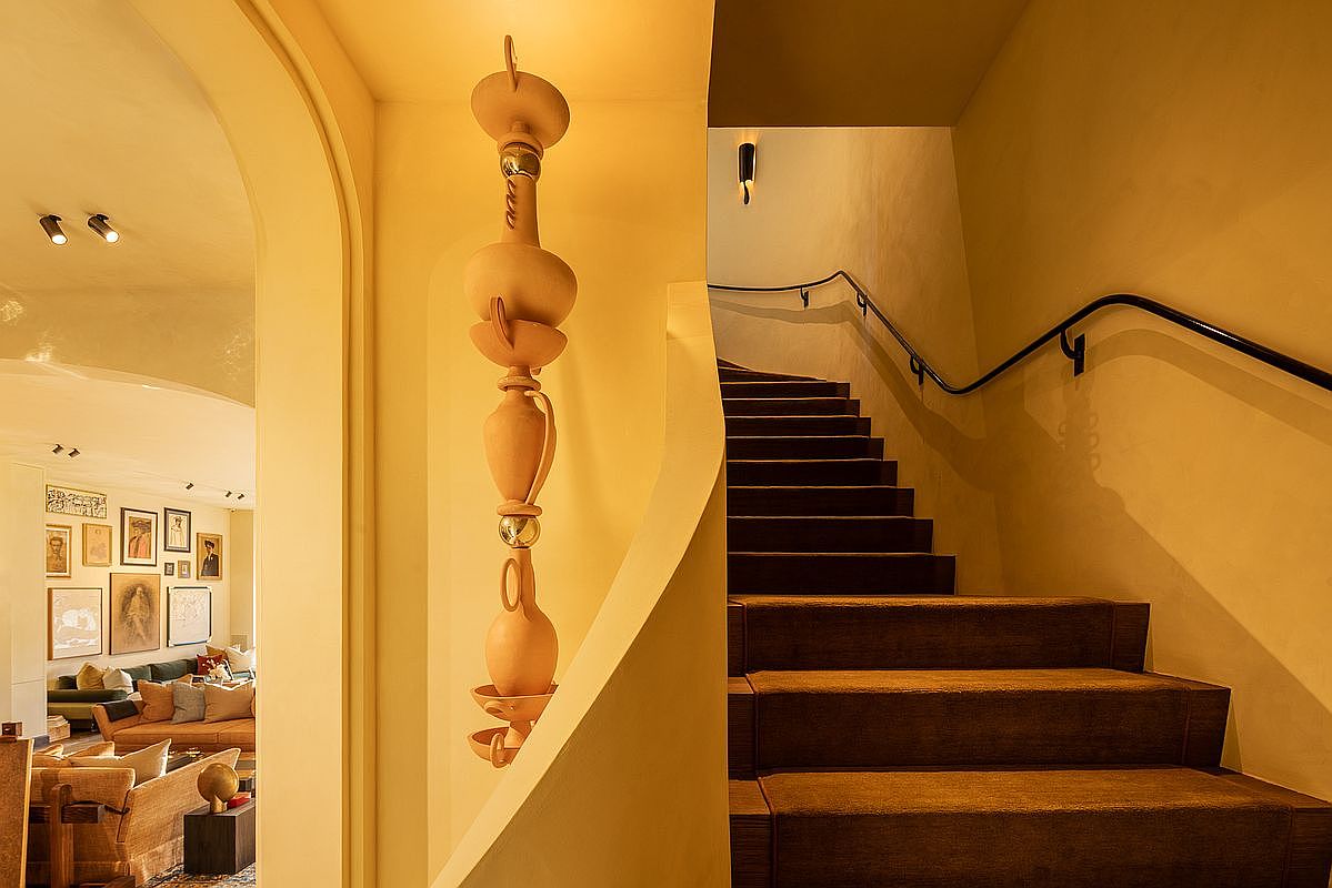 This image showcases an interior hallway and staircase, featuring warm yellow walls and a unique, sculptural light fixture. The staircase is carpeted with dark brown treads and a black handrail, leading upwards. Through an arched doorway, a glimpse of a living room with comfortable seating and artwork can be seen, creating an inviting and stylish atmosphere.