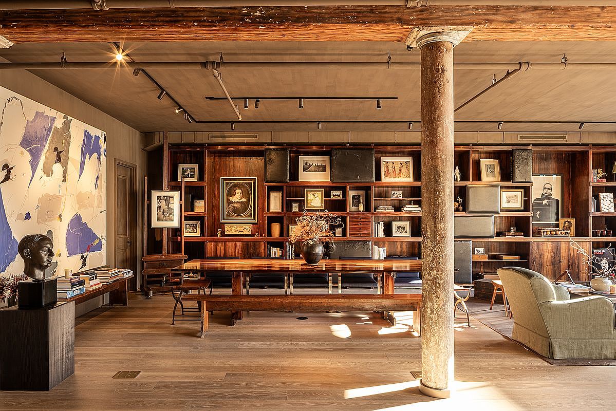 This is a spacious living room featuring a large wooden bookshelf filled with books and decorative items. A long wooden table with benches sits in the center of the room, illuminated by natural light streaming through unseen windows. The room also includes a large abstract painting and a comfortable armchair, creating a sophisticated and inviting atmosphere.