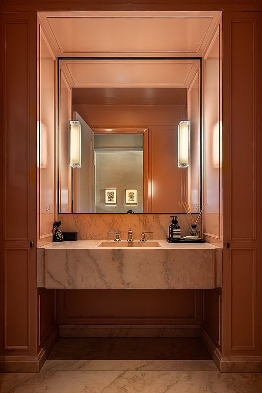 This is a well-lit bathroom featuring a marble countertop with an integrated sink and chrome fixtures. A large mirror with integrated lighting is above the sink. The walls are painted in a warm, peach tone, and the overall design is elegant and modern.