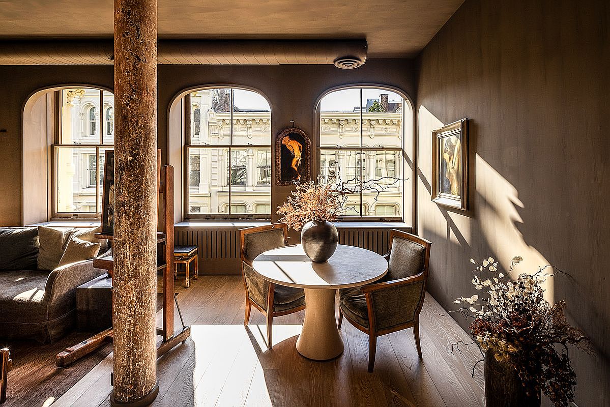 This interior shot showcases a dining area with a round table set for three, bathed in natural light streaming through arched windows. The room features hardwood floors, a decorative vase with branches on the table, and artwork on the walls, creating a warm and inviting atmosphere. A rustic wooden column adds a unique architectural element to the space.