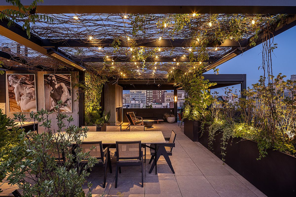 This image showcases a luxurious outdoor patio or deck area, featuring a dining table with chairs, a lounge area with comfortable seating, and lush greenery in planters. The space is illuminated by string lights overhead, creating a warm and inviting ambiance. The overall design suggests a sophisticated and relaxing outdoor living space, perfect for entertaining or enjoying quiet evenings.