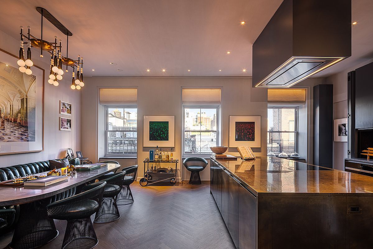 This interior shot showcases a modern kitchen and dining area with a sophisticated design. The kitchen features a large island with a metallic countertop and a sleek range hood, while the dining area includes a long table with unique wire-frame chairs and a built-in banquette. The space is well-lit with recessed lighting and a statement chandelier, creating a warm and inviting atmosphere.