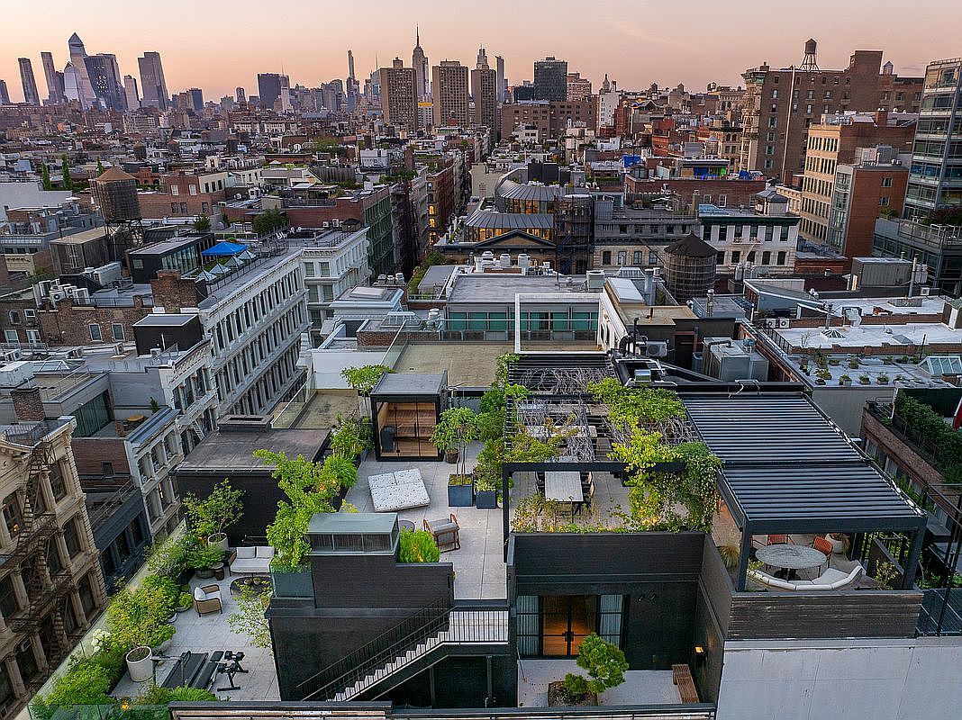 This aerial view showcases a luxurious rooftop terrace in an urban setting, complete with multiple seating areas, lush greenery, and a modern pergola. The terrace features a dining table, lounge chairs, and a covered seating area, offering various spaces for relaxation and entertainment. The backdrop includes a stunning cityscape, enhancing the property's appeal and highlighting its exclusive location.