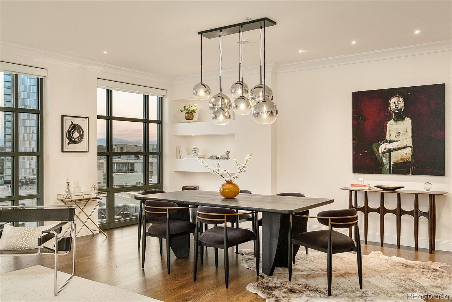 This is a well-lit dining room featuring a modern aesthetic. A large, dark-toned dining table is surrounded by six chairs with black frames and dark upholstery. An eye-catching, multi-bulb pendant light hangs above the table, and artwork adorns the walls, adding a touch of sophistication to the space.