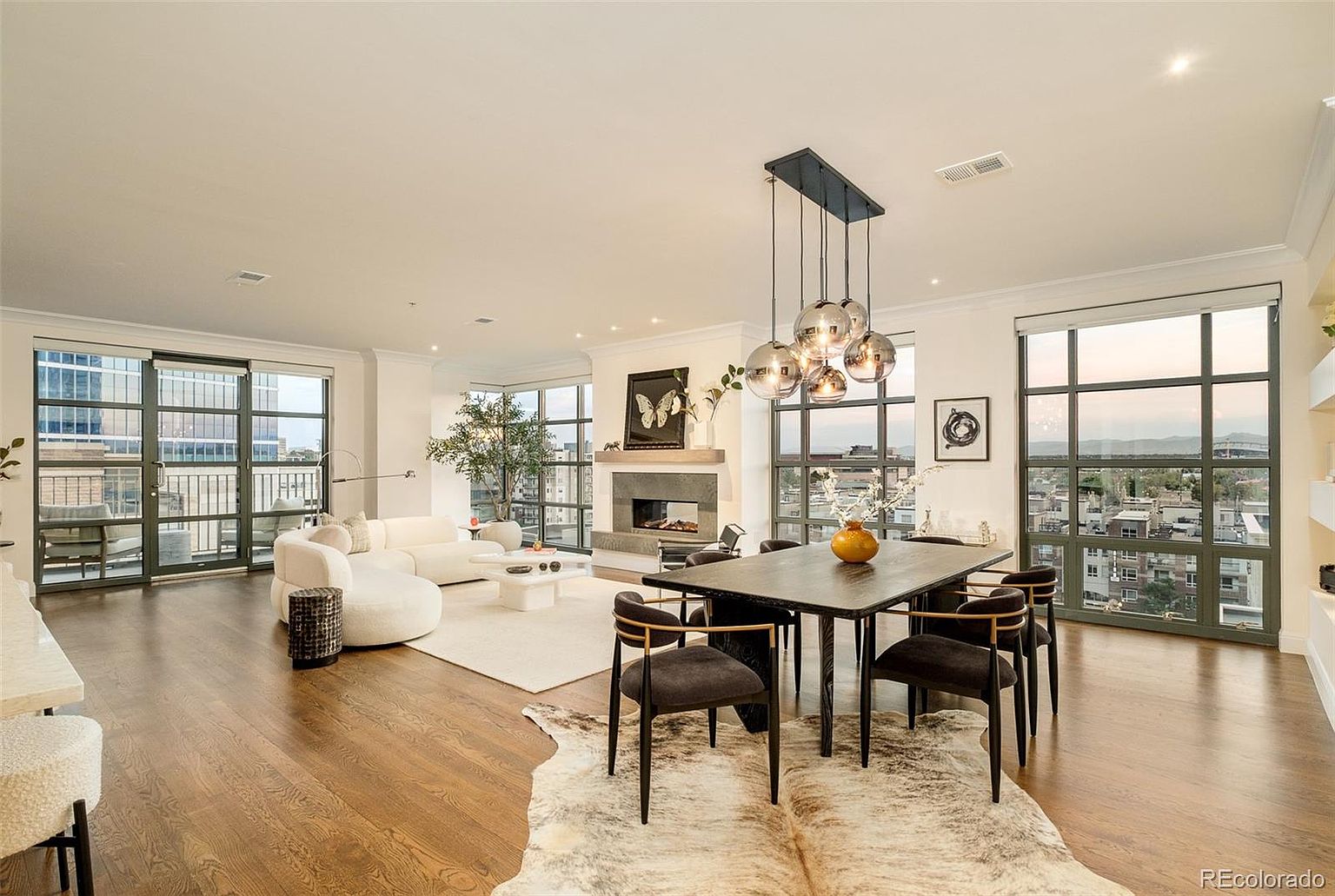 This is a spacious and luxurious living and dining area featuring hardwood floors, large windows with city views, and a modern fireplace. The room is furnished with a curved white sofa, a dark wood dining table with contemporary chairs, and a unique spherical chandelier. A cowhide rug adds texture to the dining area, while a neutral-toned area rug defines the living space.