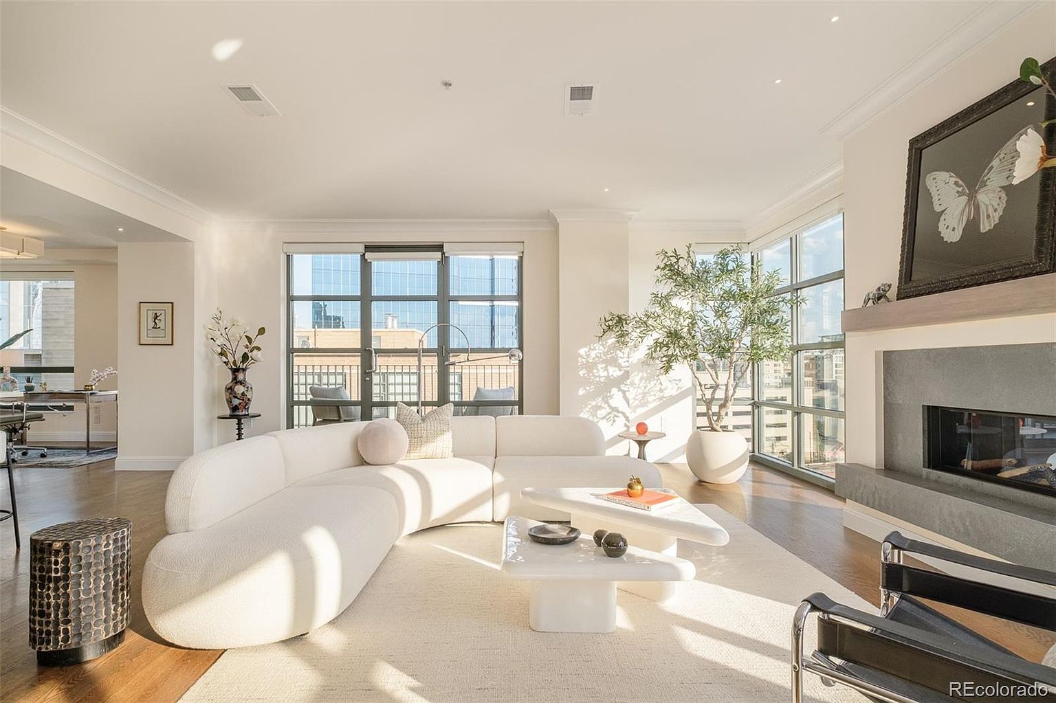 This is a bright and airy living room featuring a curved white sofa, a modern white coffee table, and a fireplace with a large framed butterfly art piece above it. Large windows provide ample natural light and views of the city. The overall aesthetic is modern and luxurious, with a focus on clean lines and neutral colors.