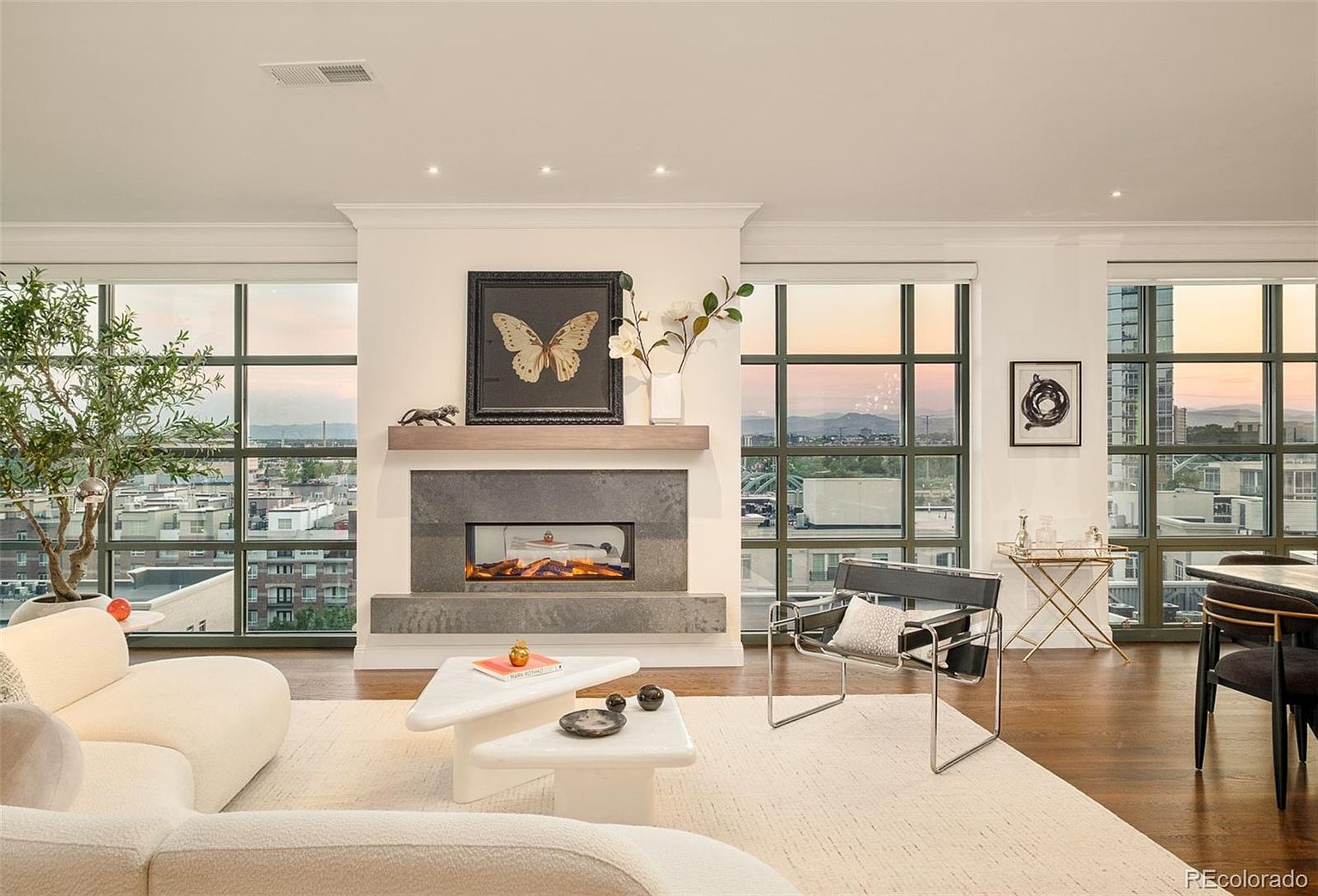 This is a bright and modern living room featuring large windows with city views, a sleek fireplace with a butterfly artwork above, and contemporary furniture including a curved sofa and a unique coffee table. The room is well-lit and airy, creating a sophisticated and inviting atmosphere. The hardwood floors and neutral color palette contribute to the room's elegant design.