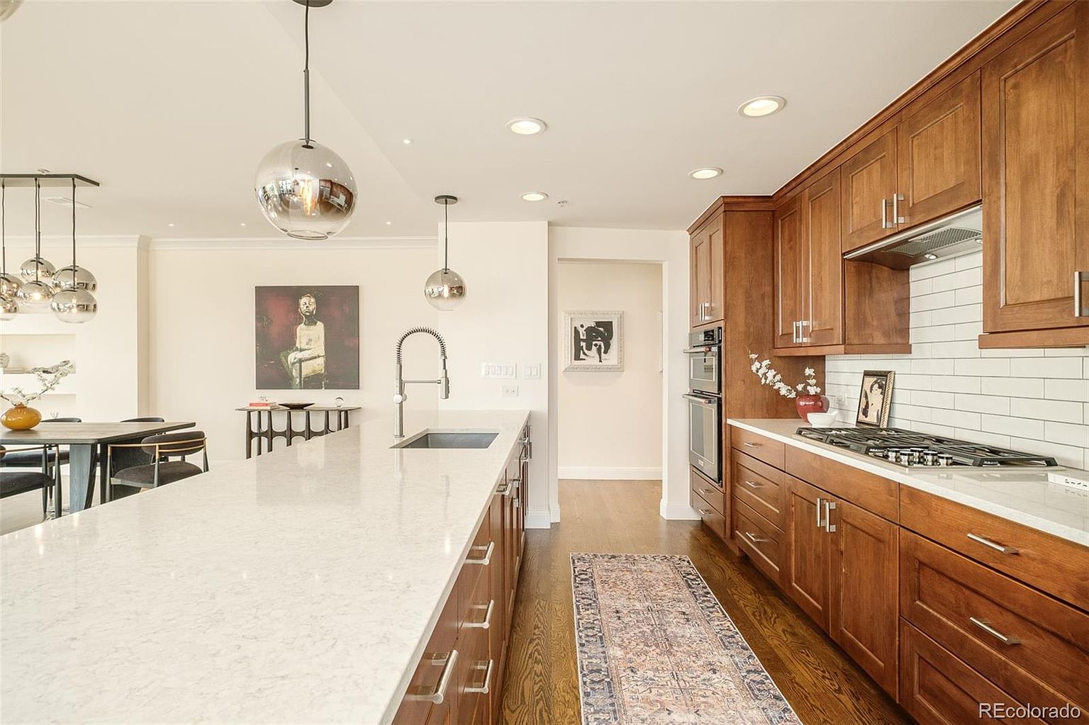 This is a well-lit kitchen featuring dark wood cabinetry, a white countertop island, and stainless steel appliances. The kitchen has a modern design with spherical pendant lights and a white subway tile backsplash. A patterned runner rug adds a touch of color to the hardwood floors.