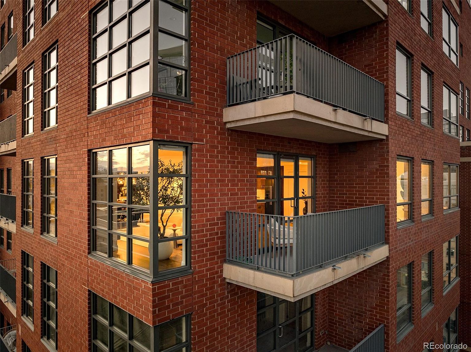This image showcases the side of a modern brick apartment building, highlighting its architectural details. The building features large windows with dark frames, offering ample natural light, and private balconies with metal railings. The warm brick tones and clean lines create a sophisticated and inviting aesthetic.