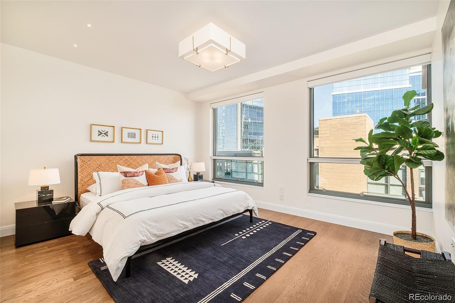 This is a well-lit primary bedroom featuring a bed with a woven headboard, white linens, and decorative pillows. The room has hardwood floors, a dark blue patterned rug, and large windows offering city views. A modern light fixture hangs from the ceiling, and there is a black nightstand with a lamp on one side of the bed.