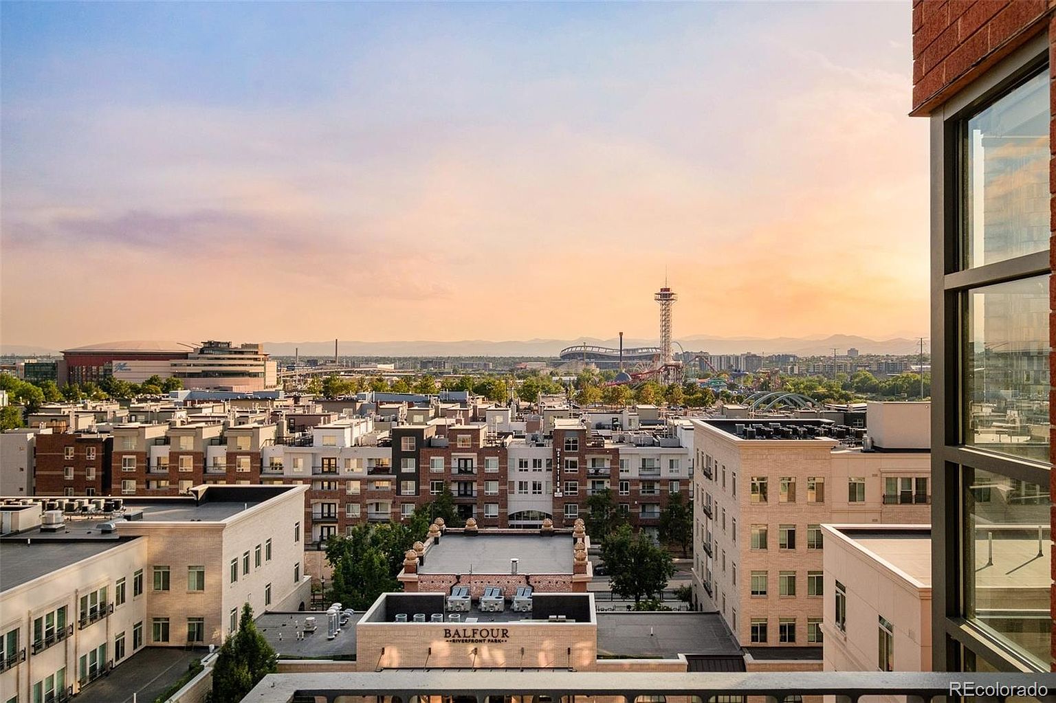 This aerial view showcases a dense urban landscape with numerous residential buildings, a stadium, and a distant mountain range under a warm, sunset-lit sky. The buildings feature a mix of architectural styles, with flat rooftops and visible balconies. The overall impression is one of a vibrant, well-established community with access to both urban amenities and natural beauty.