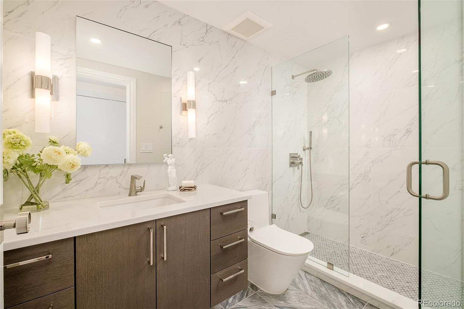 This is a well-lit primary bathroom featuring marble-like wall tiles and a modern aesthetic. The bathroom includes a vanity with a dark wood cabinet and a white countertop, a large mirror with sconces, a toilet, and a glass-enclosed shower with a rainfall showerhead. The flooring is a contrasting gray tile, and the overall impression is clean and luxurious.