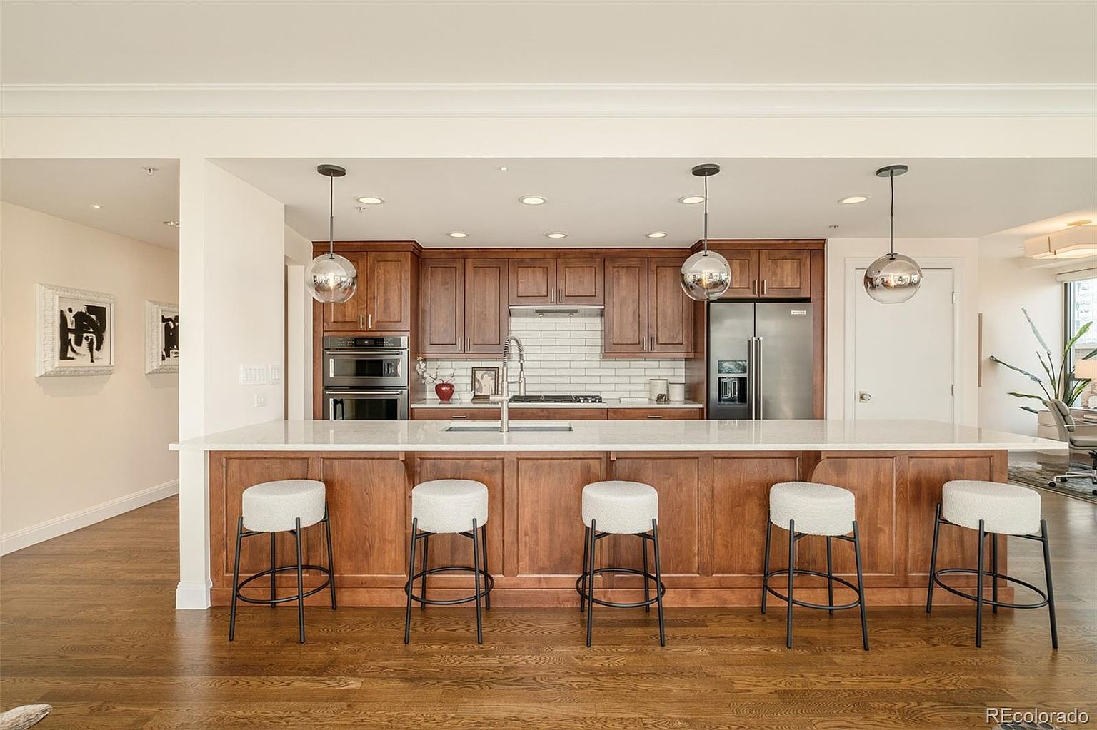 This is a well-lit kitchen featuring wooden cabinetry, stainless steel appliances, and a large island with a white countertop. Five bar stools are lined up along the island, and pendant lights hang above. The kitchen has a warm and inviting feel, with hardwood flooring adding to the overall aesthetic.