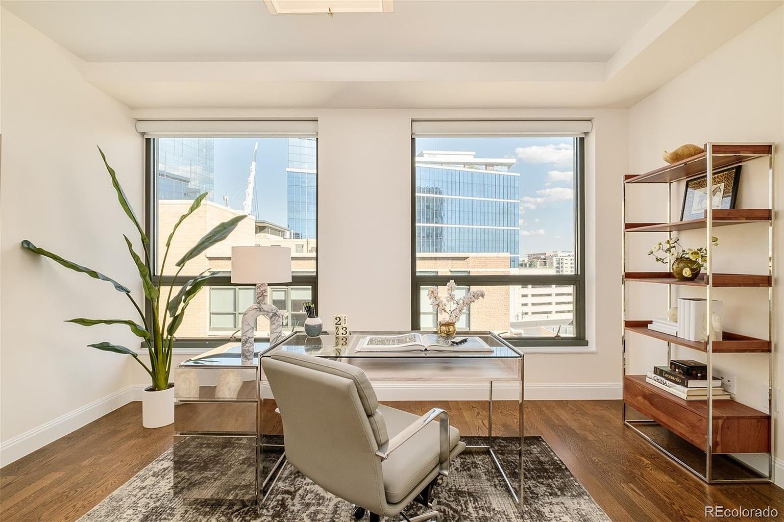 This is an interior shot of a modern office space featuring a glass-top desk with chrome accents, a gray office chair, and a tall bookshelf. Large windows provide ample natural light and views of the city skyline. A potted plant adds a touch of greenery, and a patterned rug anchors the space, creating a sophisticated and functional workspace.