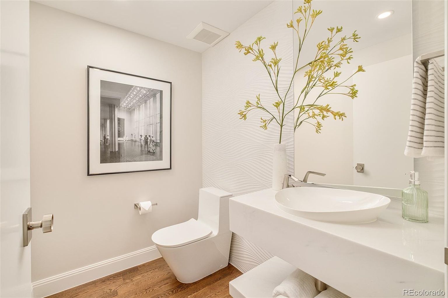 This is a modern guest bathroom featuring a floating vanity with a white countertop and an oval sink. The walls are painted in a neutral tone, complemented by a textured accent wall. A black and white framed print hangs above the toilet, and the flooring is a warm wood tone, creating a stylish and inviting space.