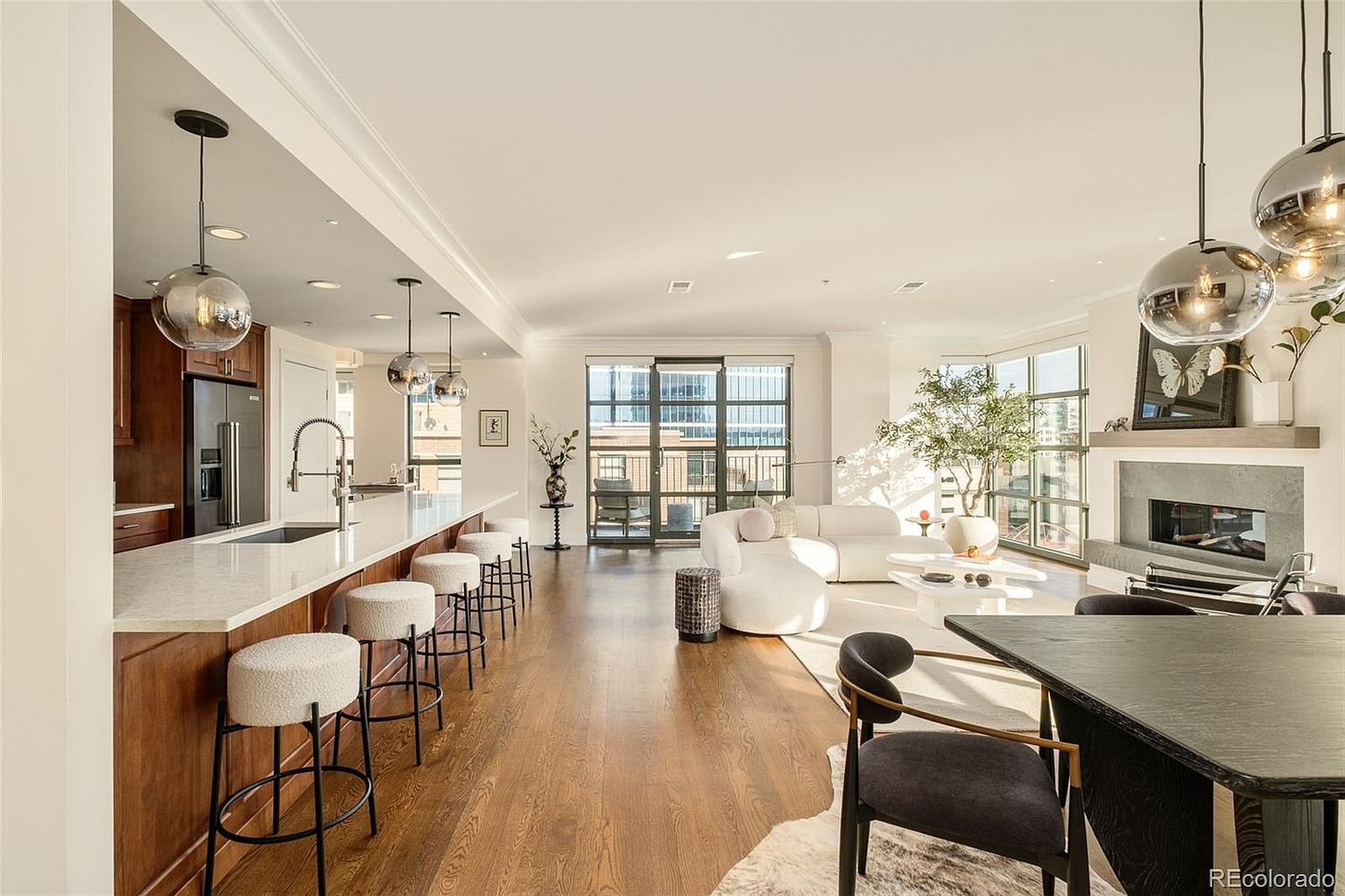 This is a wide-angle shot of a modern living space that seamlessly integrates a kitchen and dining area. The kitchen features a long island with bar stools and pendant lighting, while the living area boasts a fireplace, large windows, and contemporary furniture. The hardwood floors and neutral color palette create a bright and inviting atmosphere.