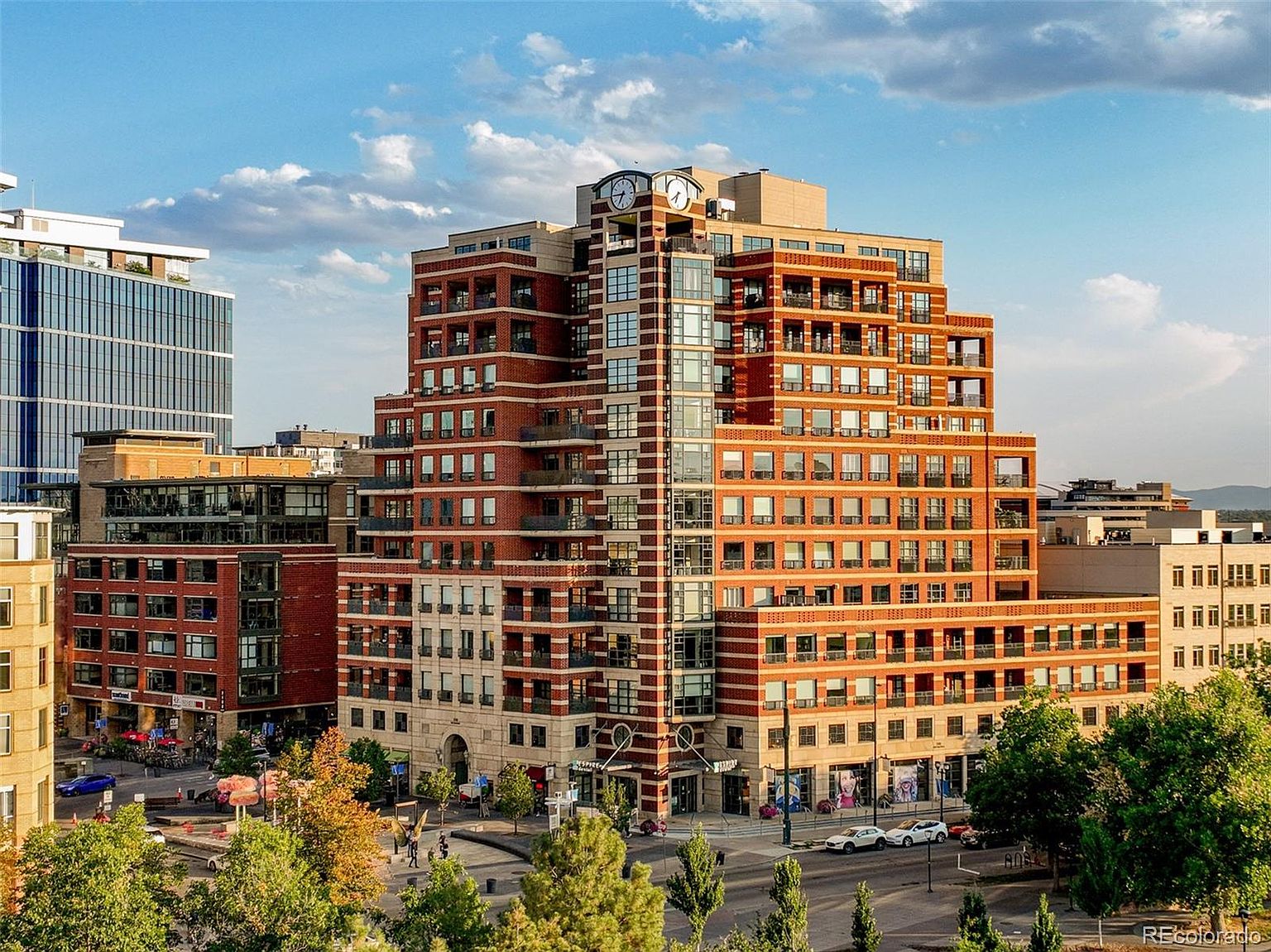This is a front view of a multi-story brick building with a clock tower. The building features a mix of brick and glass, with balconies on many floors. The surrounding area includes trees, streets, and other buildings, creating a vibrant urban setting.