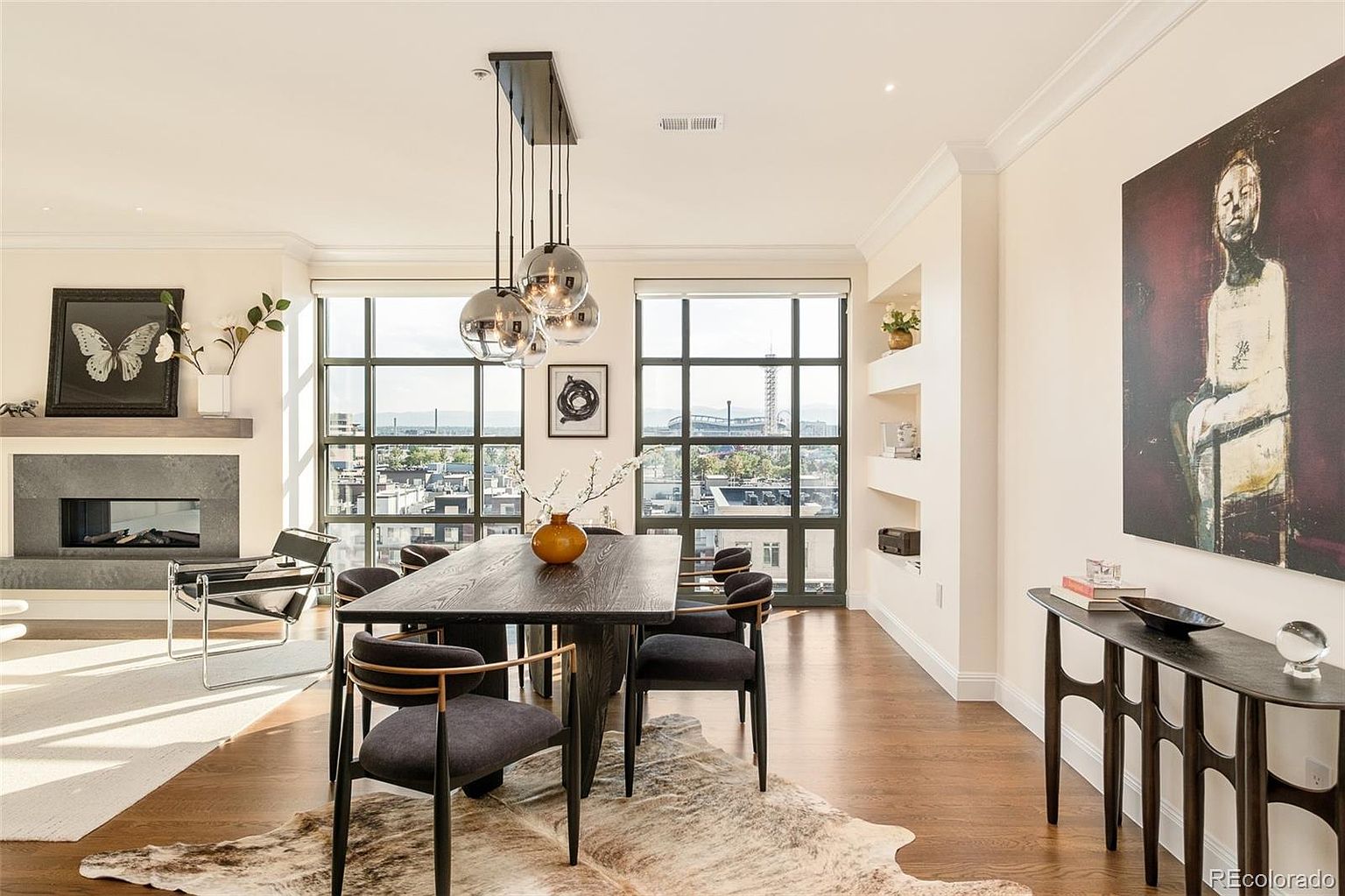 This interior shot showcases a stylish dining room with a long, dark wood table surrounded by modern chairs with gray cushions and gold accents. A unique chandelier with multiple glass globes hangs above the table, and large windows offer a city view. The room also features a fireplace, a console table with artwork, and a cowhide rug, creating a sophisticated and inviting atmosphere.