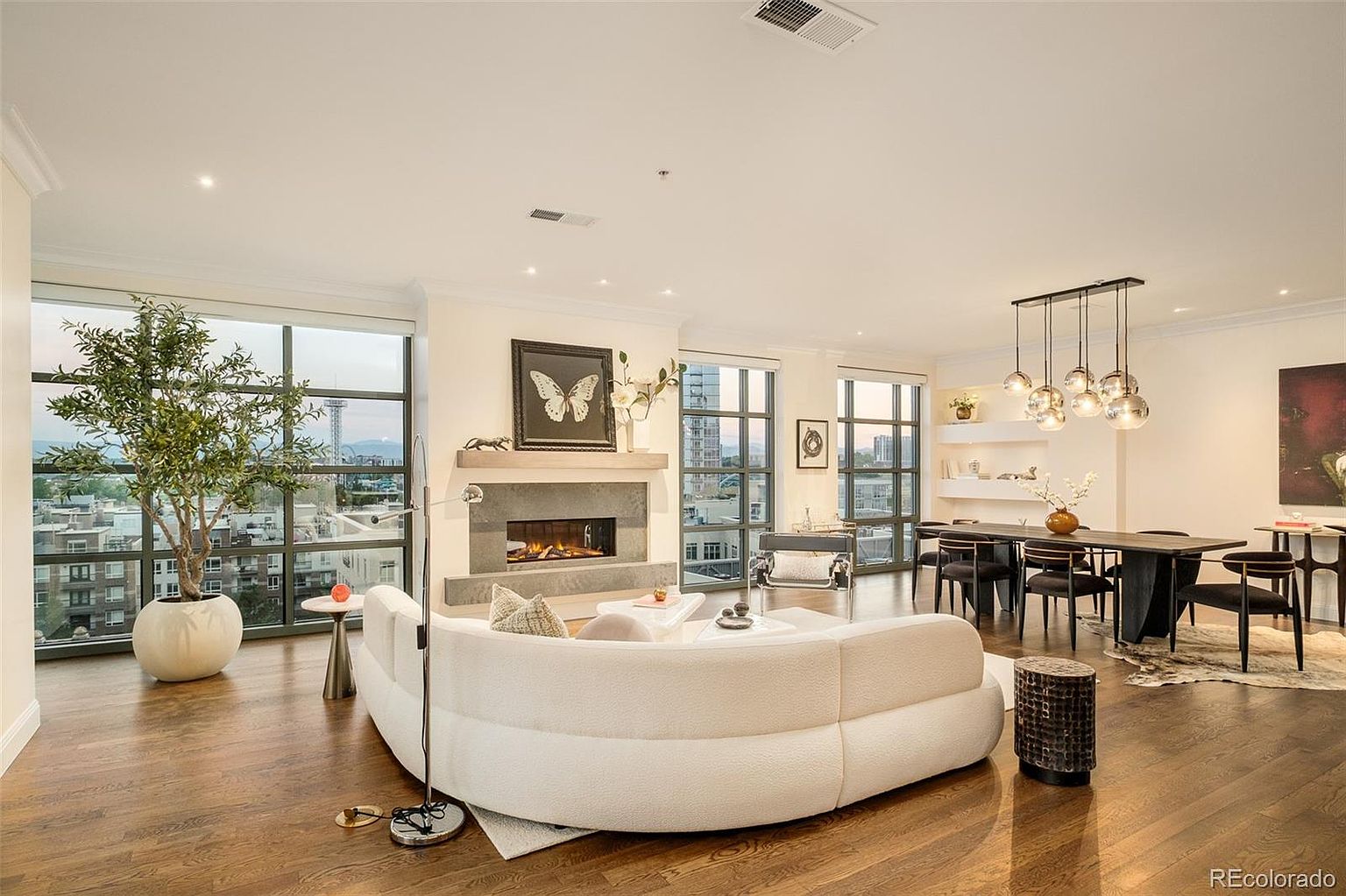 This is a spacious and modern living room featuring hardwood floors, large windows with city views, and a unique curved sofa. A fireplace adds a cozy touch, while the open layout seamlessly connects to the dining area. The room is well-lit and exudes a sophisticated and comfortable ambiance.