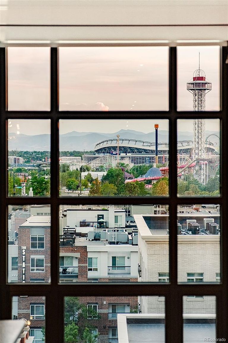 This image showcases a view from a window, framing a cityscape that includes a stadium, amusement park rides, and surrounding buildings. The window's grid pattern creates distinct visual sections, highlighting different aspects of the urban landscape and distant mountains. The perspective offers a unique vantage point, blending indoor comfort with an expansive outdoor scene.