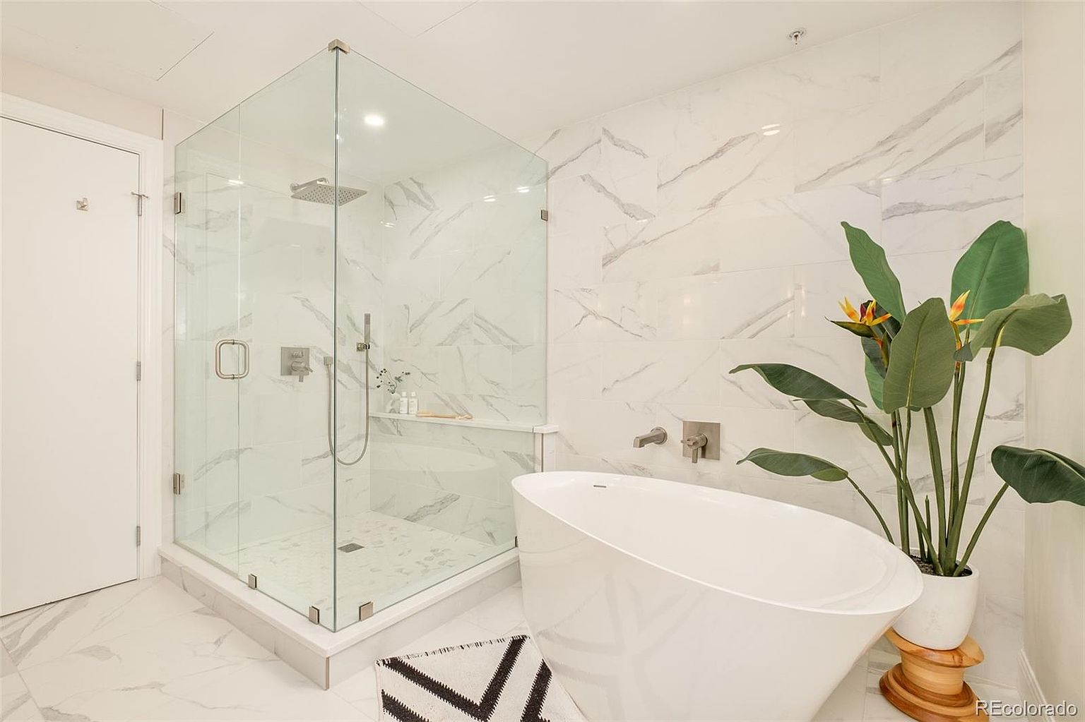 This is a bright and modern primary bathroom featuring a glass-enclosed shower with marble-patterned walls and a sleek, freestanding bathtub. A large potted plant adds a touch of nature to the space, which is finished with matching marble flooring and minimalist fixtures. The overall impression is clean, luxurious, and spa-like.