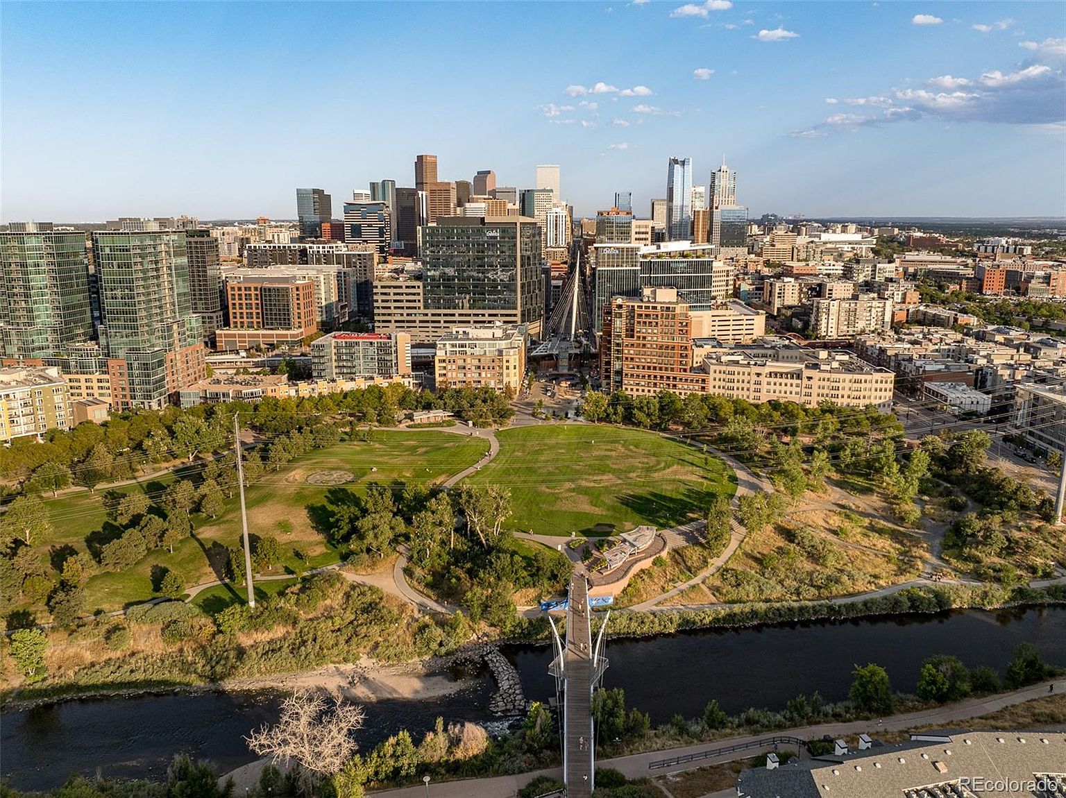 This aerial view showcases a vibrant cityscape with modern buildings and lush green spaces. A river flows through the foreground, crossed by pedestrian bridges, leading the eye towards a large park. The overall impression is one of a thriving urban environment with ample recreational opportunities.