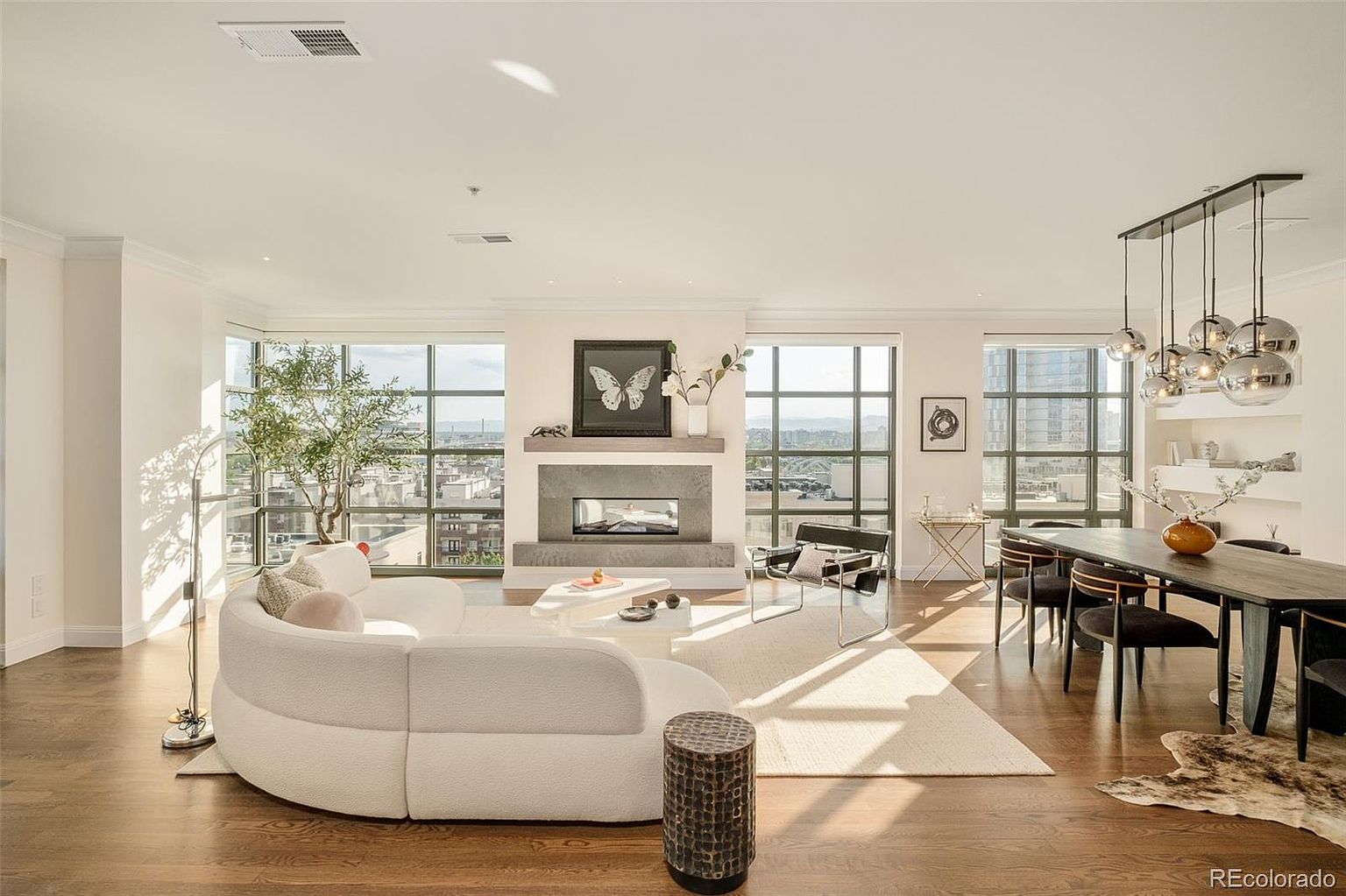 This is a bright and airy living and dining area featuring hardwood floors, large windows with city views, and a modern fireplace. The living room is furnished with a curved white sofa and a unique side table, while the dining area includes a dark wood table with upholstered chairs and a contemporary chandelier. The overall impression is one of sophisticated urban living.