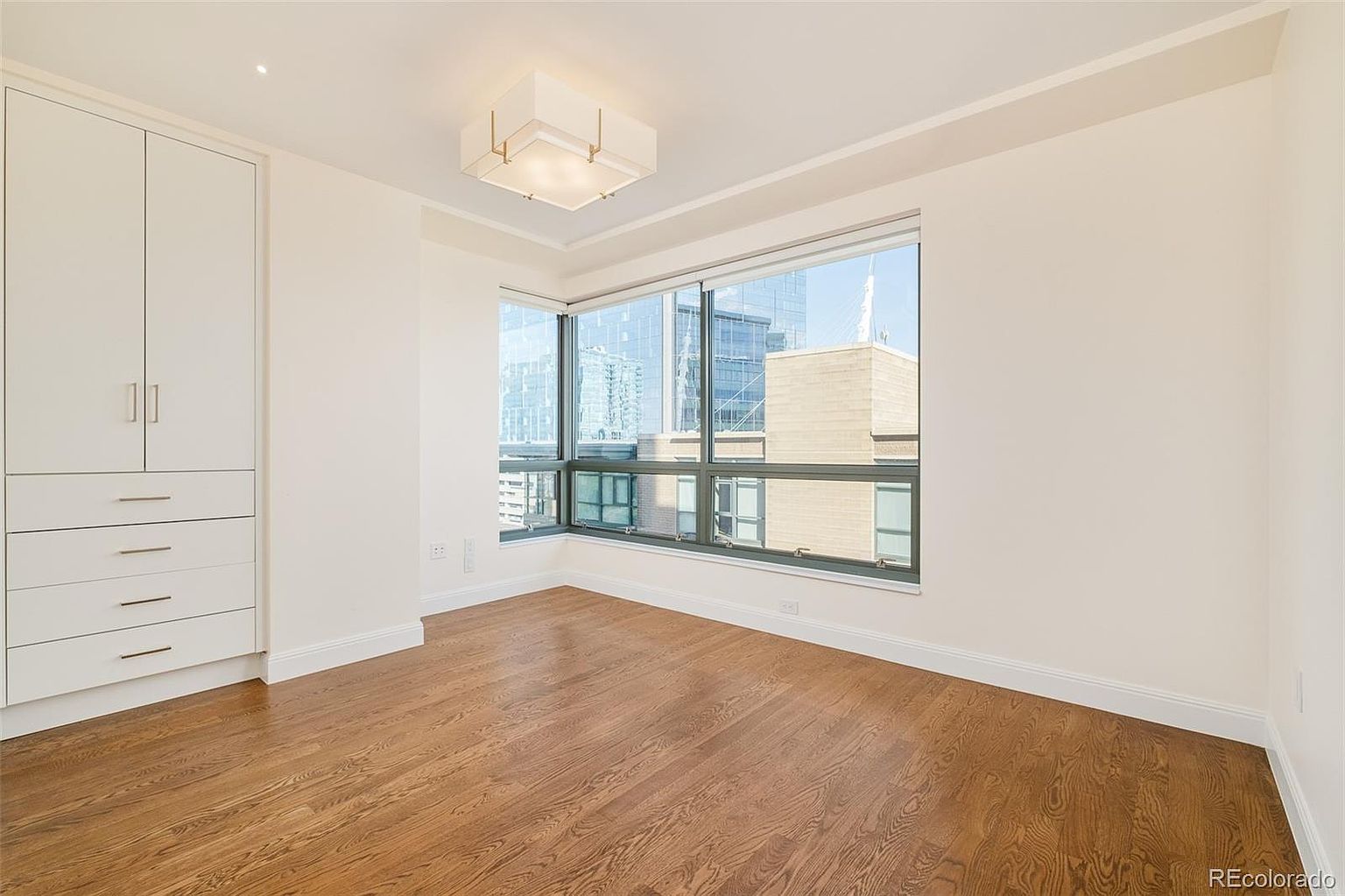 This is a bright and airy bedroom featuring hardwood floors and a large window offering city views. A built-in wardrobe provides ample storage space. The room is painted in neutral tones, creating a calm and inviting atmosphere, perfect for a guest room or secondary bedroom.