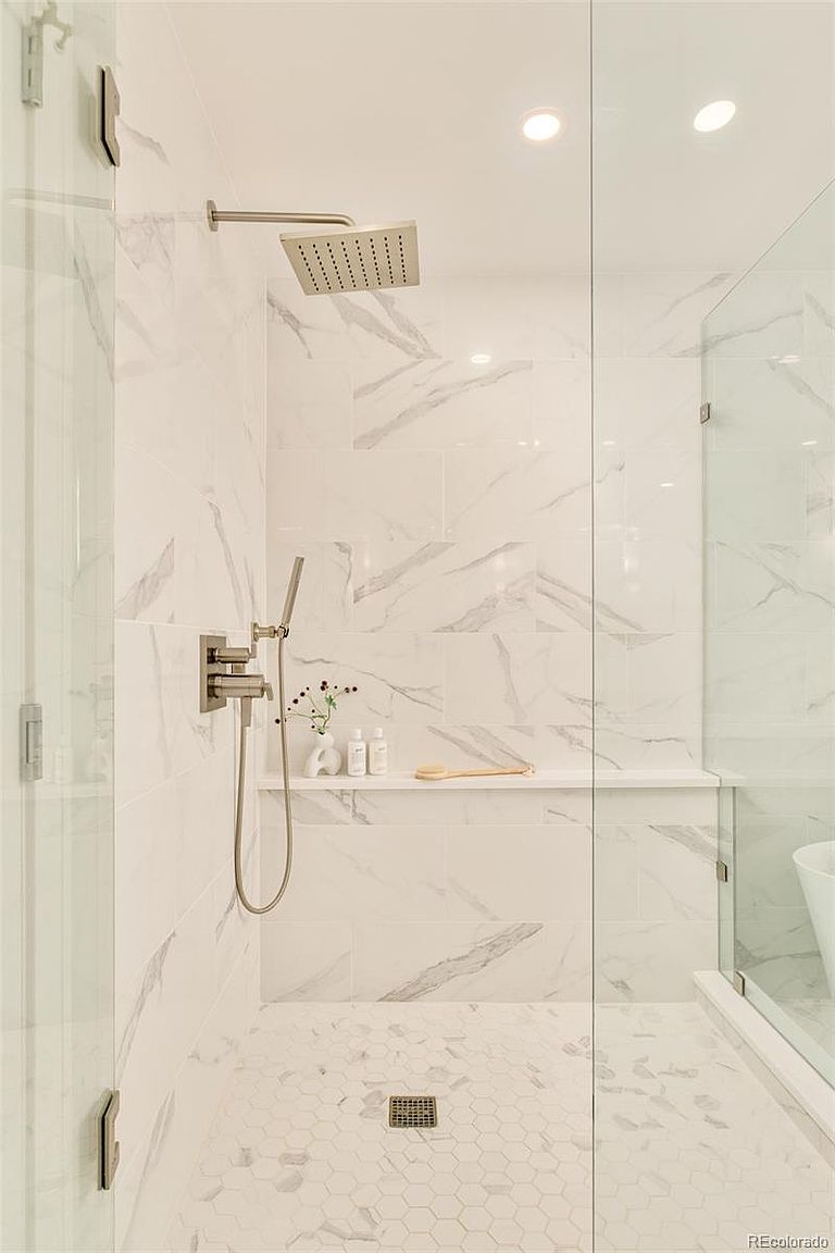 This is a well-lit primary bathroom featuring a modern shower with white marble-patterned walls and hexagonal floor tiles. The shower includes a rainfall showerhead, a handheld shower wand, and a built-in shelf for toiletries. The space is enclosed with clear glass panels, creating a clean and luxurious feel.