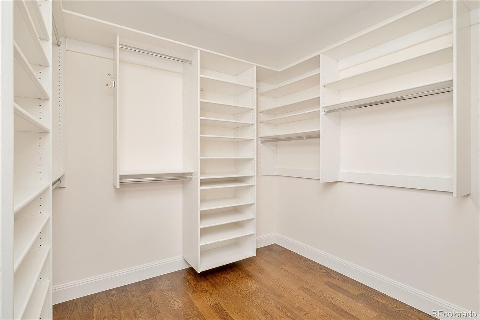 This is a well-organized walk-in closet featuring a custom shelving system with multiple shelves and hanging rods. The closet is painted in a neutral color, and the hardwood flooring adds a touch of elegance. The space appears clean and spacious, offering ample storage.