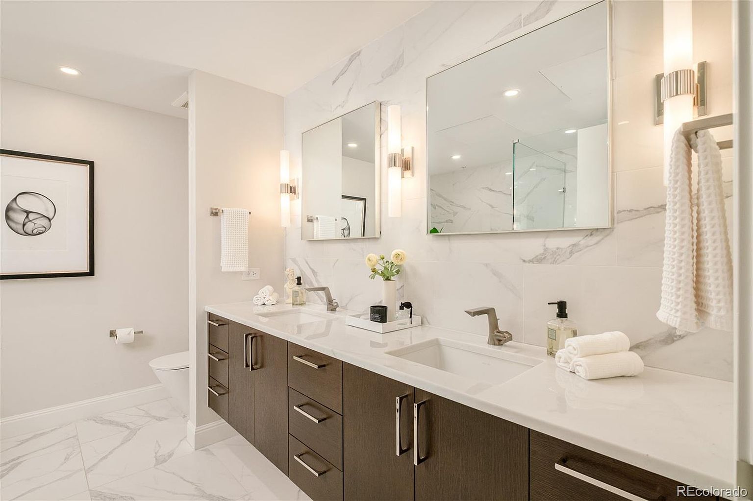 This is a primary bathroom featuring a double vanity with dark wood cabinets and white countertops. Two square mirrors are mounted above the sinks, flanked by modern sconces. The walls and floors are covered in white marble, creating a bright and luxurious space.
