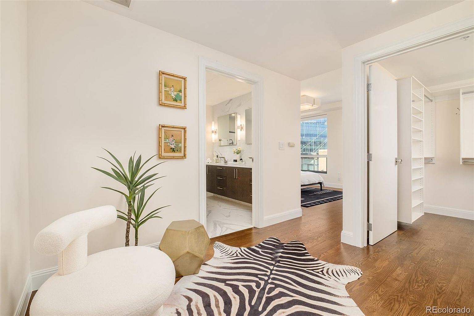 This interior shot showcases a stylish hallway leading to various rooms. The hallway features hardwood flooring, white walls adorned with framed artwork, and a zebra-print rug adding a touch of modern flair. Open doorways reveal glimpses of a bathroom, bedroom, and walk-in closet, creating an inviting and well-connected living space.