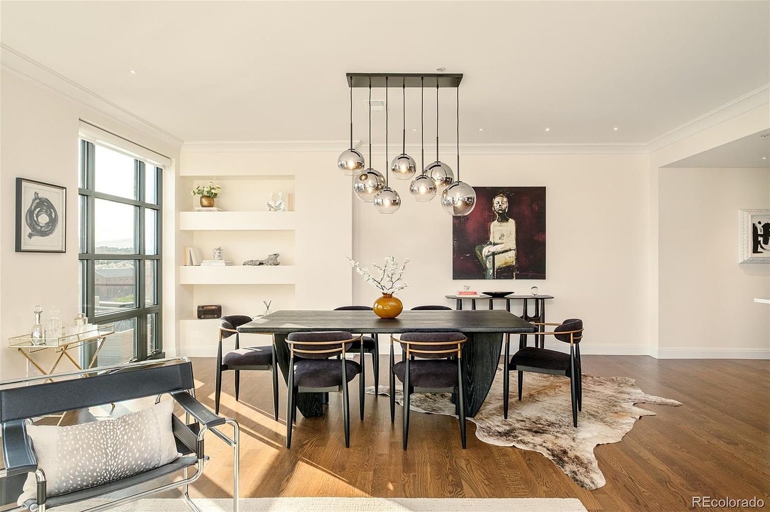 This is an interior shot of a dining room featuring a dark wood table surrounded by six chairs with black upholstery and gold accents. A modern chandelier with multiple glass globes hangs above the table, and a large painting adorns the wall. The room has hardwood floors and a neutral color palette, creating a sophisticated and inviting atmosphere.