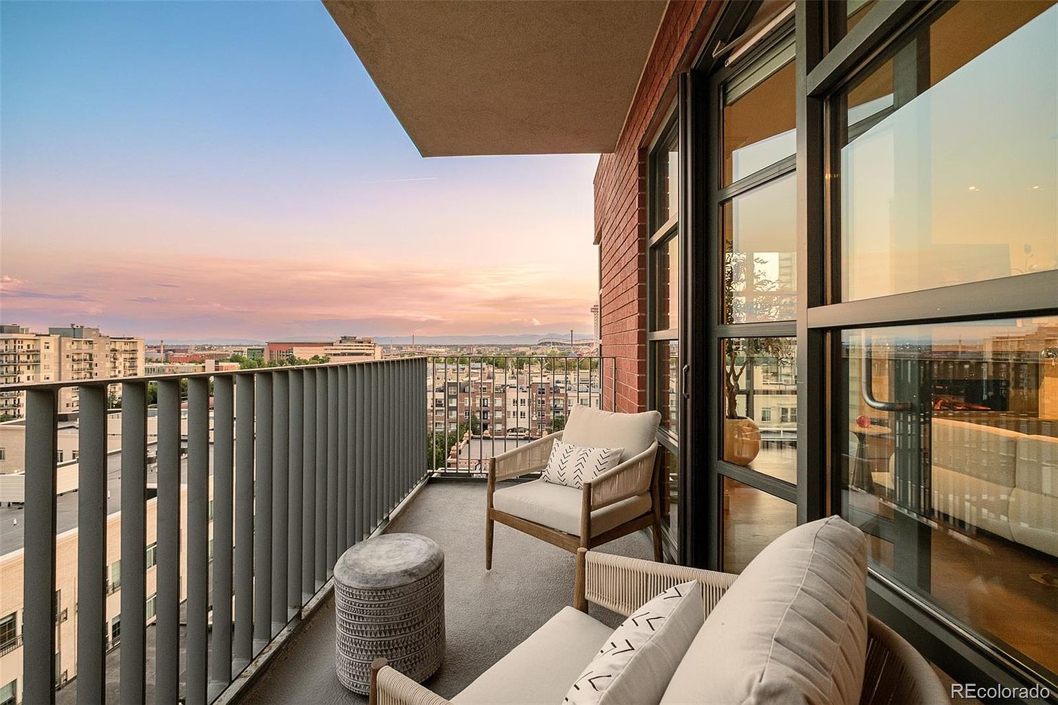 This image showcases a stylish balcony with comfortable seating, including a chair and a cushioned bench, complemented by a textured ottoman. The balcony features a modern railing and offers a scenic city view under a vibrant sunset sky. Large glass doors provide seamless indoor-outdoor living, enhancing the appeal of this urban residence.