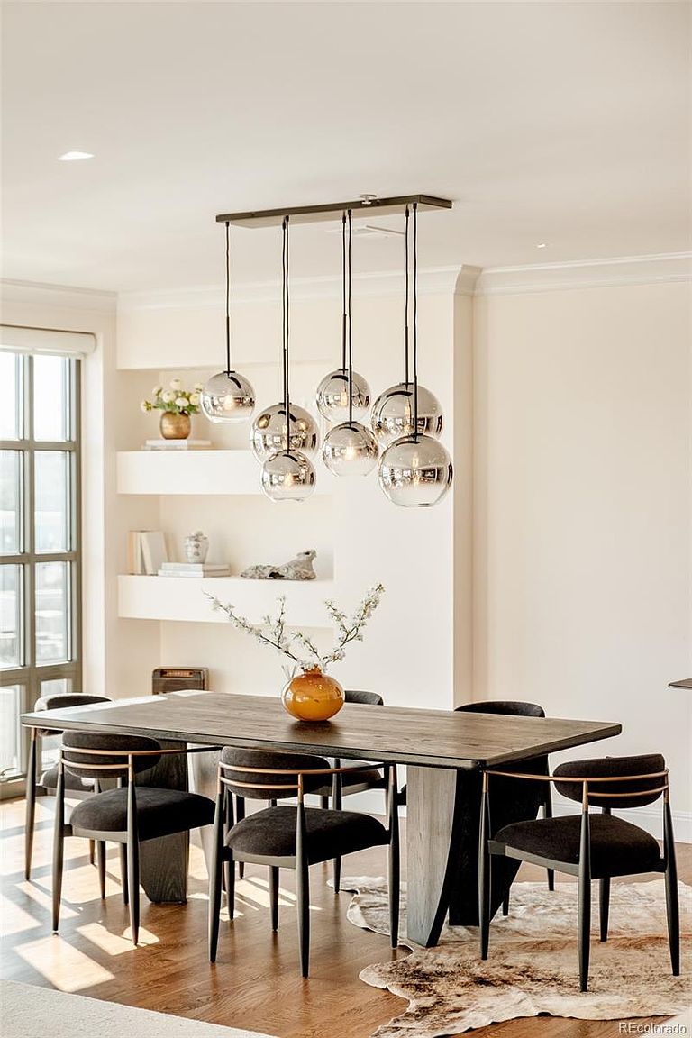 This is an interior shot of a dining room featuring a long, dark wood table with a light-colored base, surrounded by black chairs with gold accents. An arrangement of spherical pendant lights hangs above the table, and a vase with branches sits in the center. The room has a modern and elegant feel, with neutral wall colors and built-in shelving in the background.