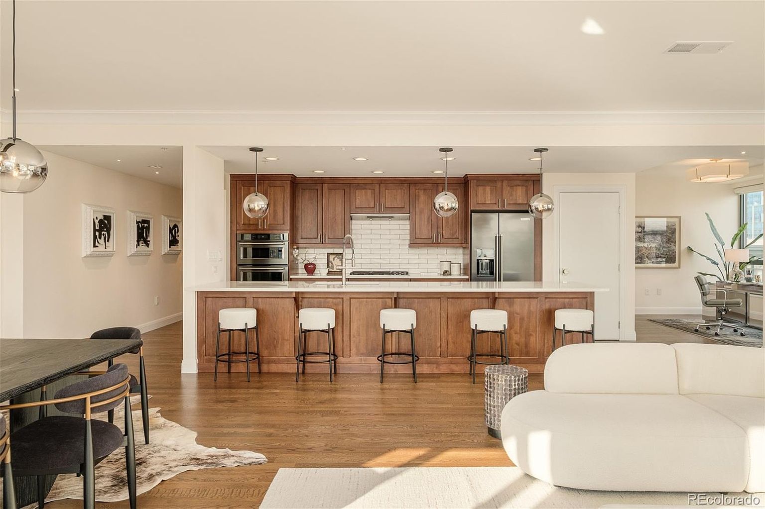 This interior shot showcases a modern kitchen with rich wood cabinetry and a white countertop island, complemented by stylish pendant lighting. The kitchen seamlessly blends into the living space, featuring a dining area with contemporary chairs and a comfortable white sofa, creating an open and inviting atmosphere. Natural light floods the space, highlighting the hardwood floors and enhancing the overall warmth of the home.
