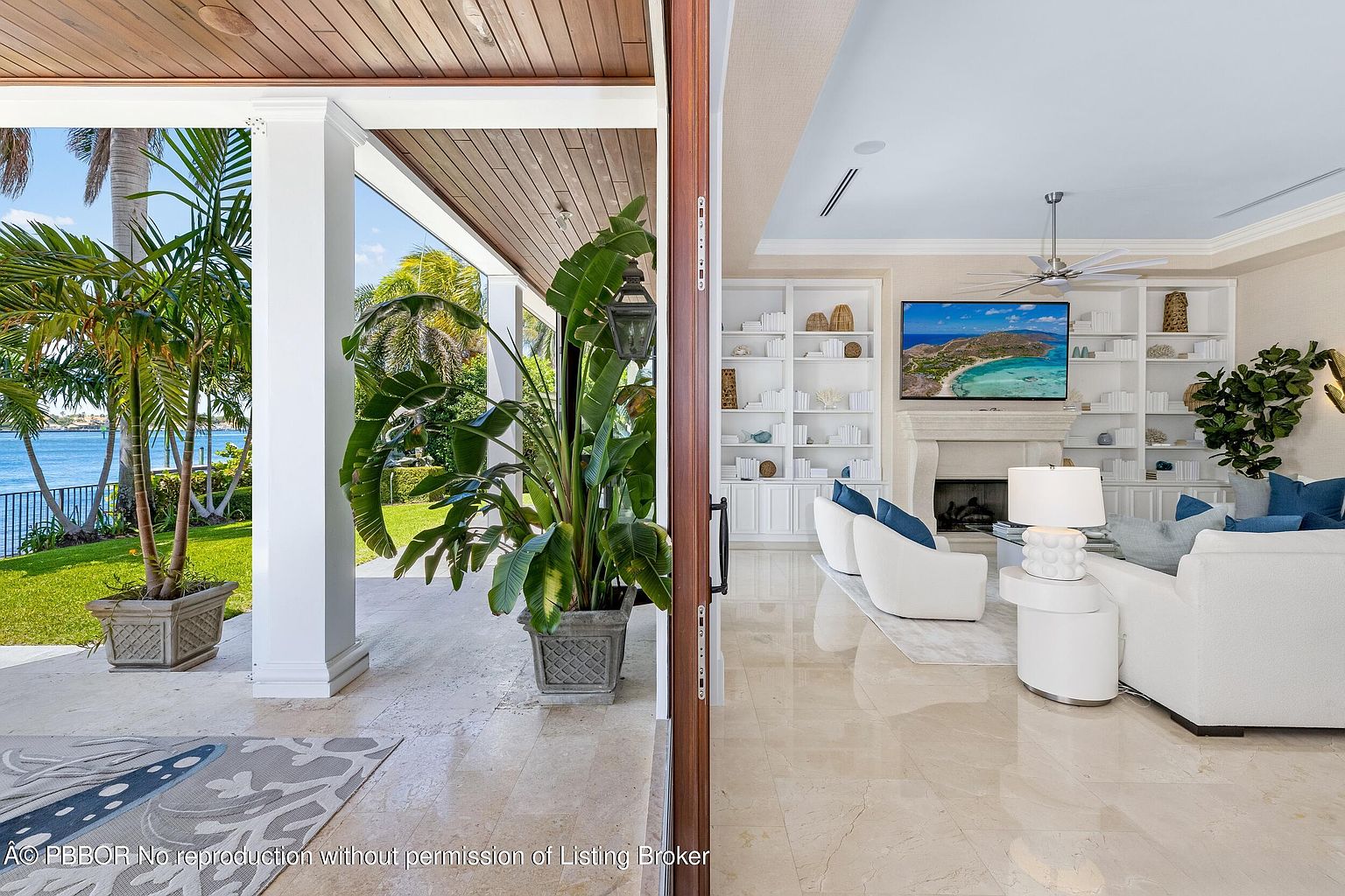 This image showcases a luxurious living room viewed through an open doorway, blending indoor and outdoor elements. The interior features a bright, elegant living space with white furniture, built-in bookshelves, and a fireplace with a television above. Outside, a covered patio with lush greenery and a glimpse of the waterfront create a seamless transition between the interior and the natural surroundings.