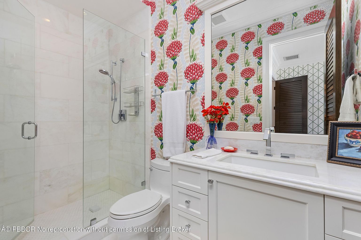 This is a well-lit bathroom featuring a glass-enclosed shower with white marble-like tiles and a white toilet. The vanity has a white cabinet with a marble countertop and a white sink. The walls are decorated with a red floral wallpaper, adding a pop of color to the space.
