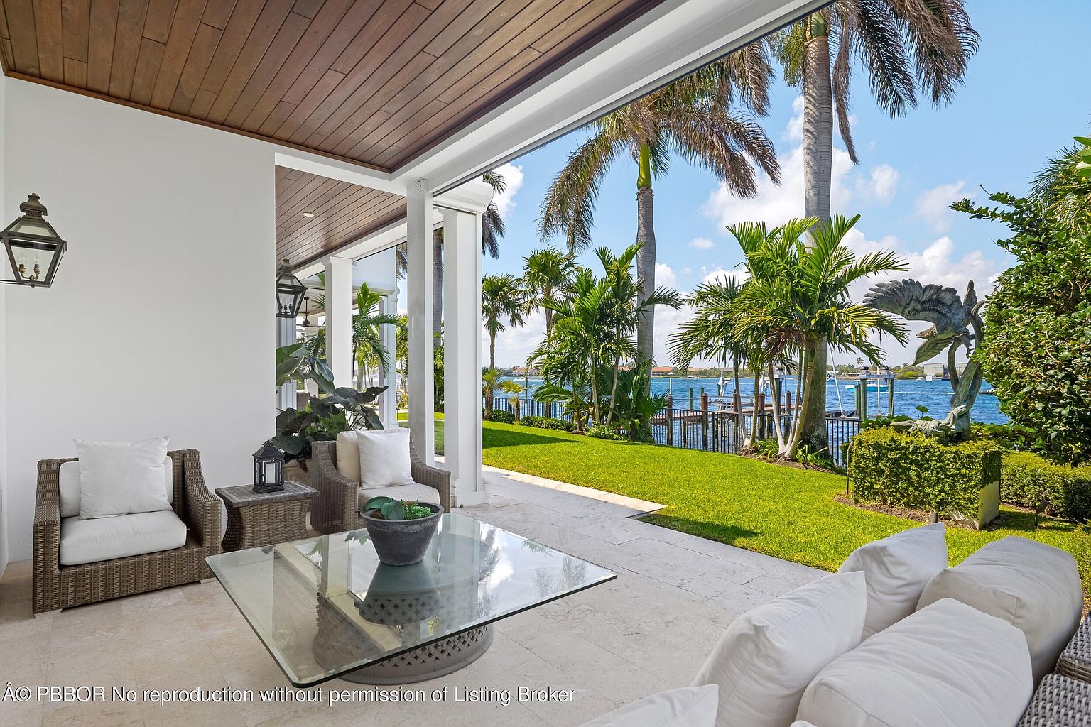 This image showcases a luxurious outdoor patio area with comfortable wicker seating and a glass-topped coffee table. The space is partially covered by a wooden ceiling and features white support columns, offering a seamless transition to the lush green lawn and waterfront view with palm trees. The overall impression is one of relaxed elegance and tropical sophistication.
