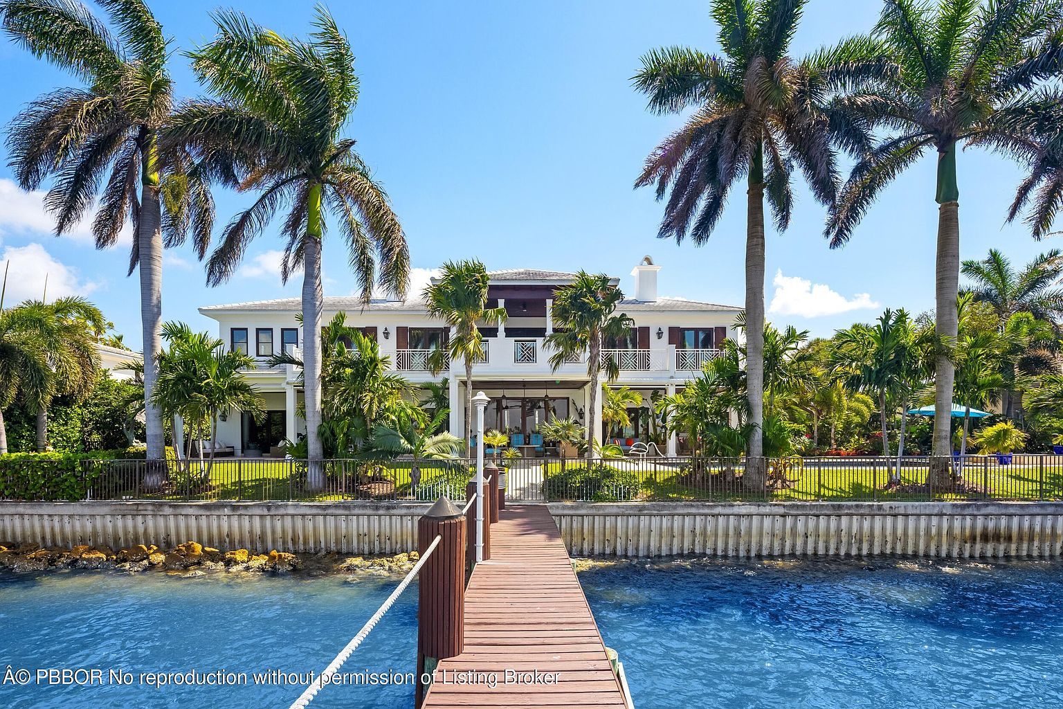 This image showcases the rear exterior of a luxurious waterfront property, framed by tall palm trees and lush landscaping. A private wooden dock extends into the clear blue water, offering direct access for boating or swimming. The house features multiple balconies and a well-manicured lawn, creating an inviting and serene atmosphere.