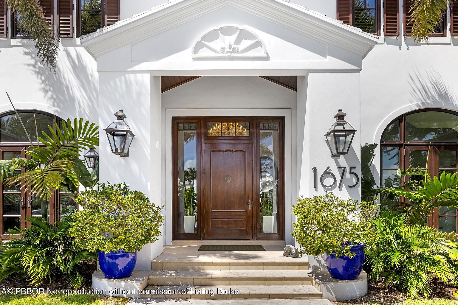 This is a welcoming entryway to a luxurious home. The focal point is a grand wooden door flanked by glass panels and illuminated by a decorative transom. Flanking the steps are vibrant blue planters filled with lush greenery, and elegant lanterns adorn the walls, adding to the sophisticated ambiance. The house number '1675' is prominently displayed, and the overall impression is one of refined elegance and curb appeal.