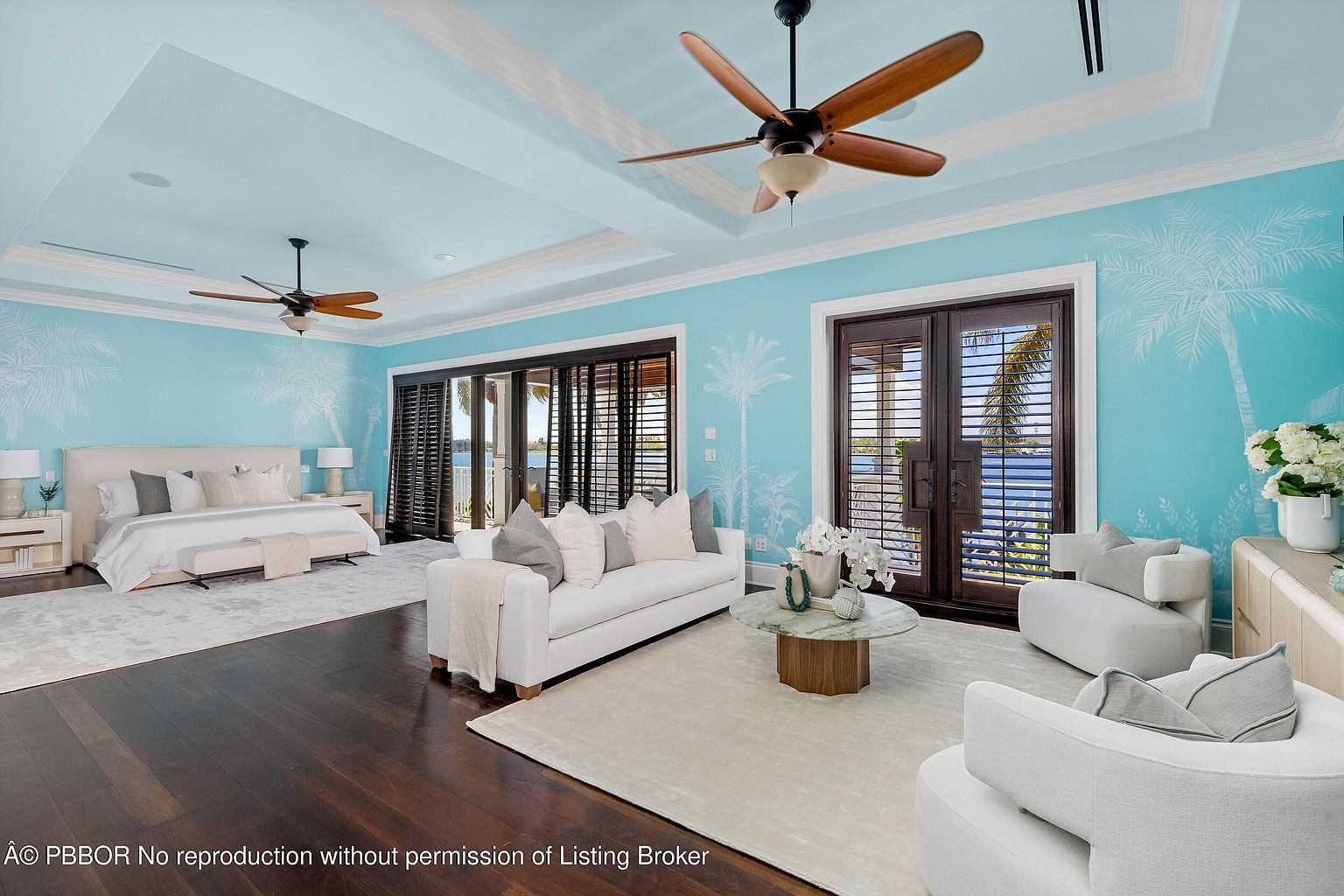 This is a primary bedroom featuring a king-size bed with neutral bedding, a seating area with a white sofa and armchairs, and access to a balcony with water views. The walls are painted a light blue with palm tree accents, and the room is well-lit with natural light and ceiling fans. The overall style is luxurious and coastal.