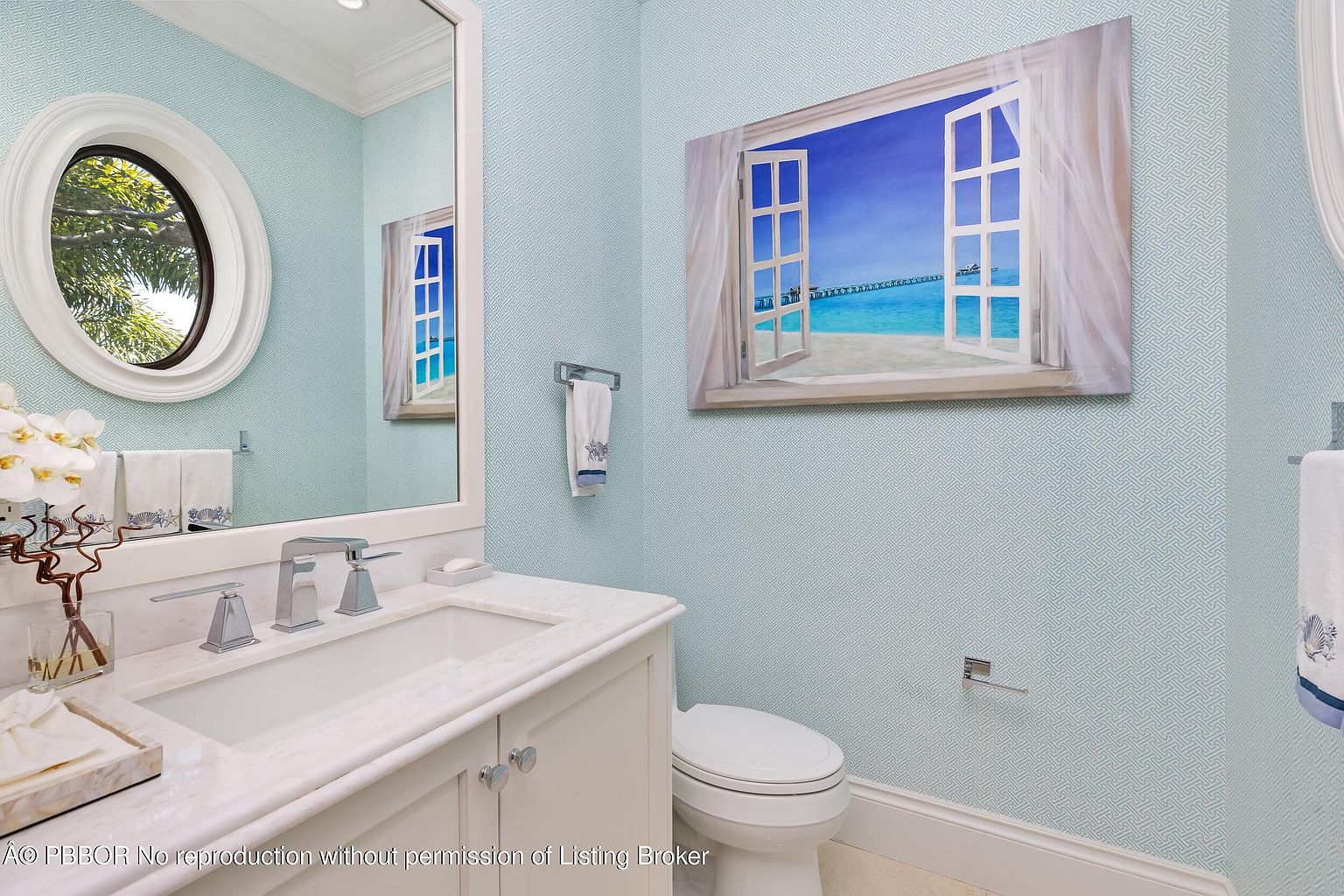 This is a well-lit guest bathroom featuring a vanity with a white marble countertop and a white cabinet. The walls are covered in a light blue patterned wallpaper, and a framed painting of a beach scene hangs above the toilet. A round mirror and decorative towels add to the room's coastal aesthetic.
